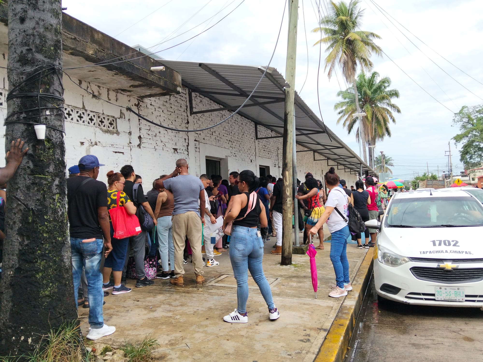 People stand on a sidewalk in line next to a white brick building
