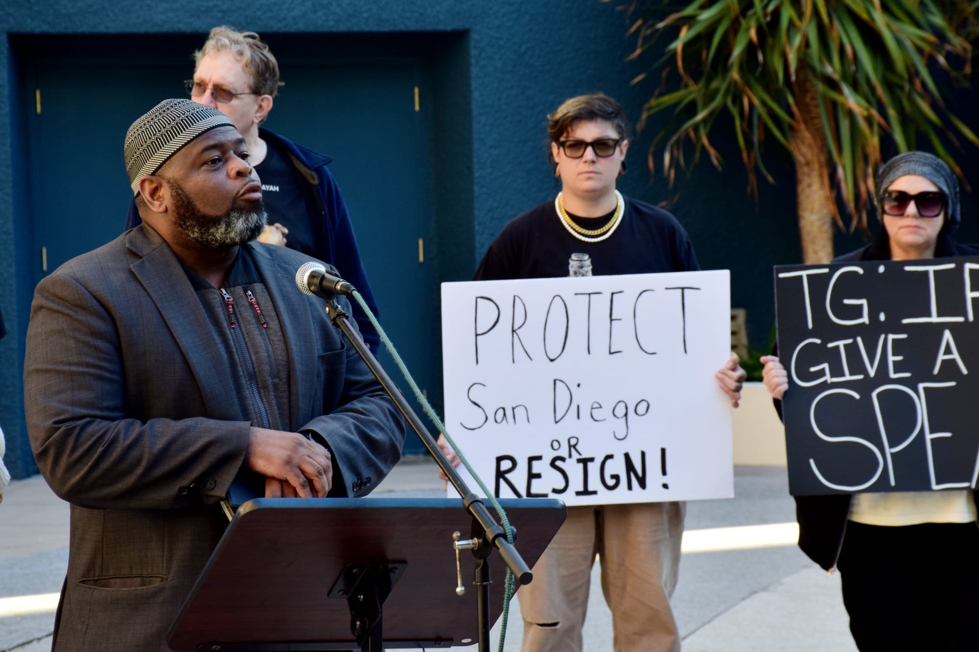 A man speaks at a podium outside, with several people standing and holding signs behind him.
