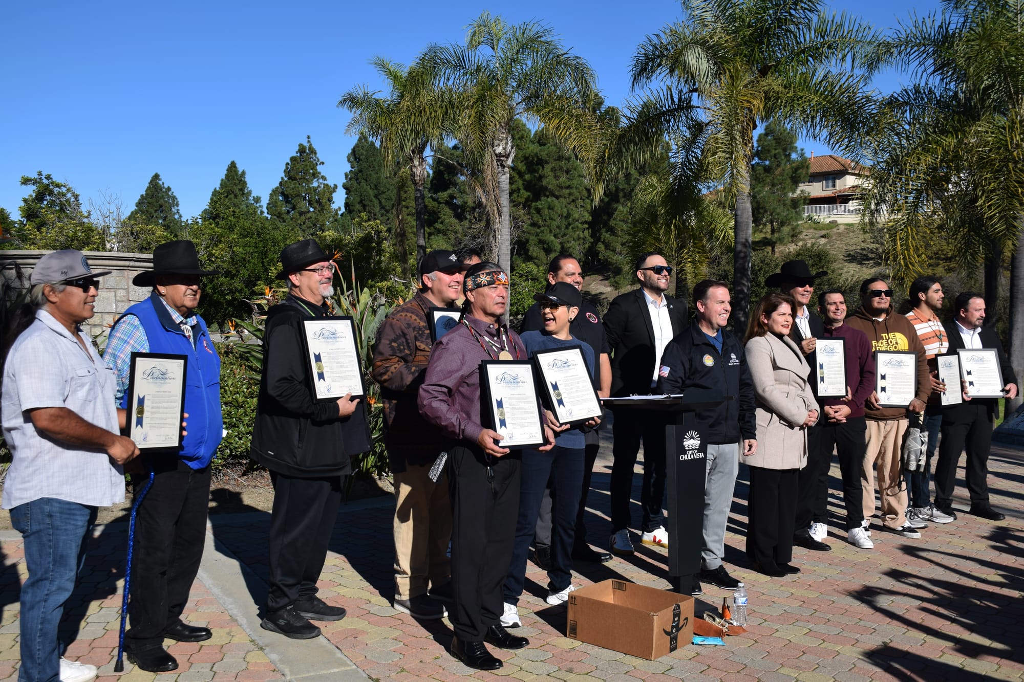 A group of people stand in a line holding framed copies of a proclamation outside in front of palm trees 