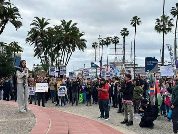 A woman wearing a hijab and light-colored clothing uses a microphone to address a crowd of people holding signs with palm trees in the background
