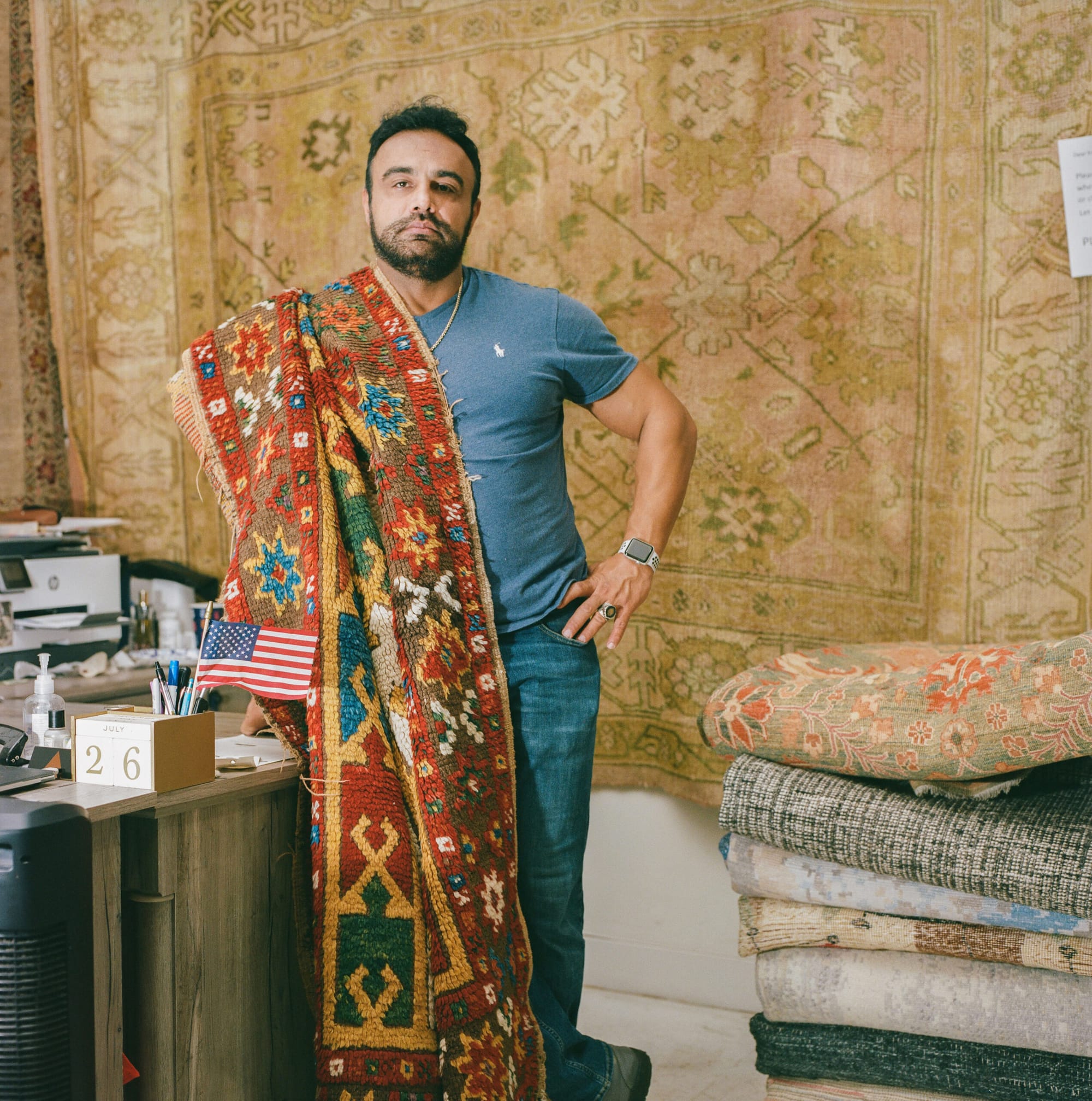 A man stands in front of rugs with another rug draped over his shoulder and the American flag on a desk next to him