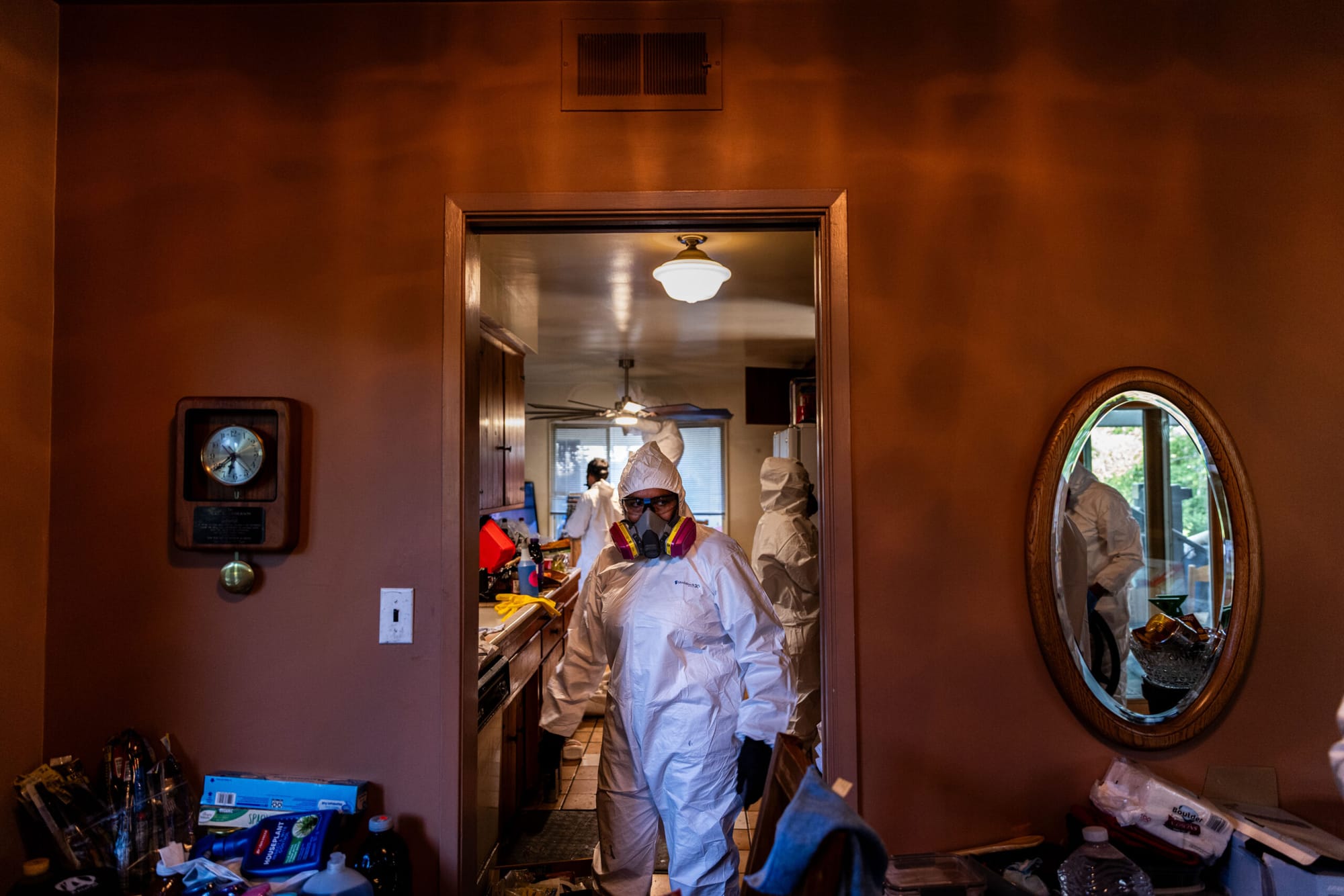 A person in personal protective equipment stands in a doorway with other people in ppe behind her cleaning a kitchen