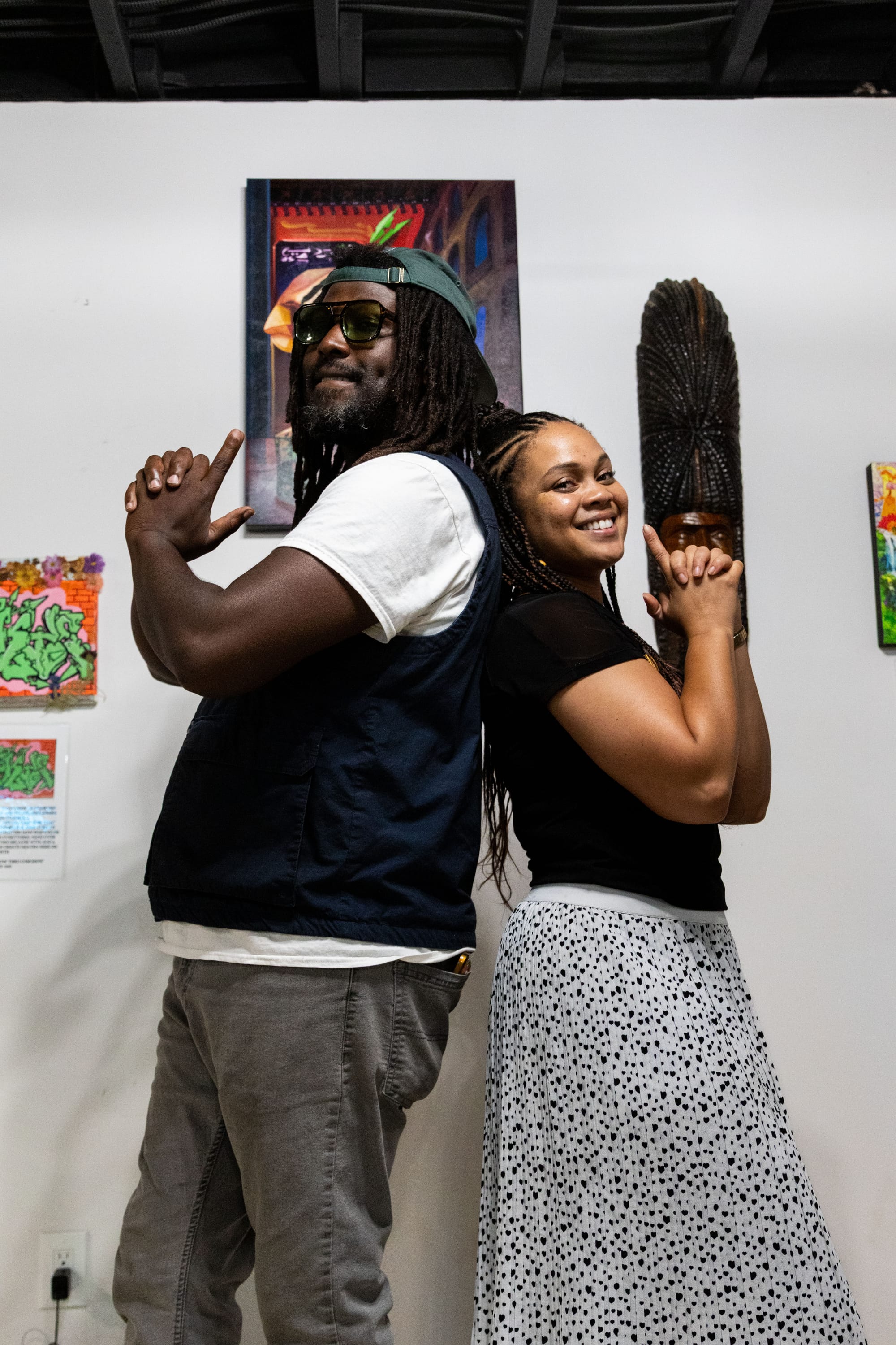 Two people stand back to back posting for a photograph in front of a gallery wall.
