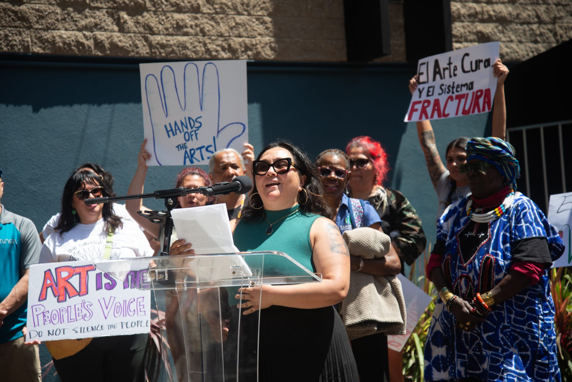 A woman in a green top and black skirt speaks at a podium surrounded by people with colorful signs