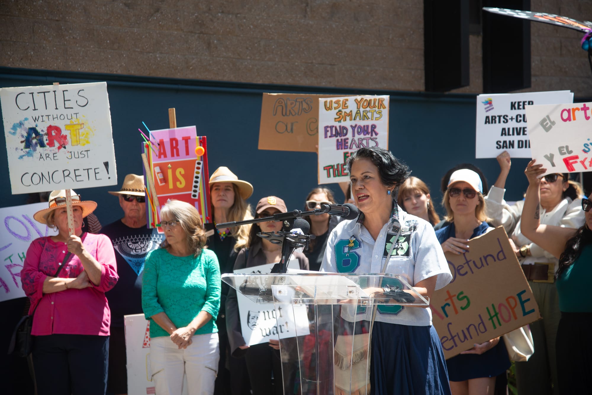 A woman in a blue skirt and light-colored top stands at a podium surrounded by people with colorful signs