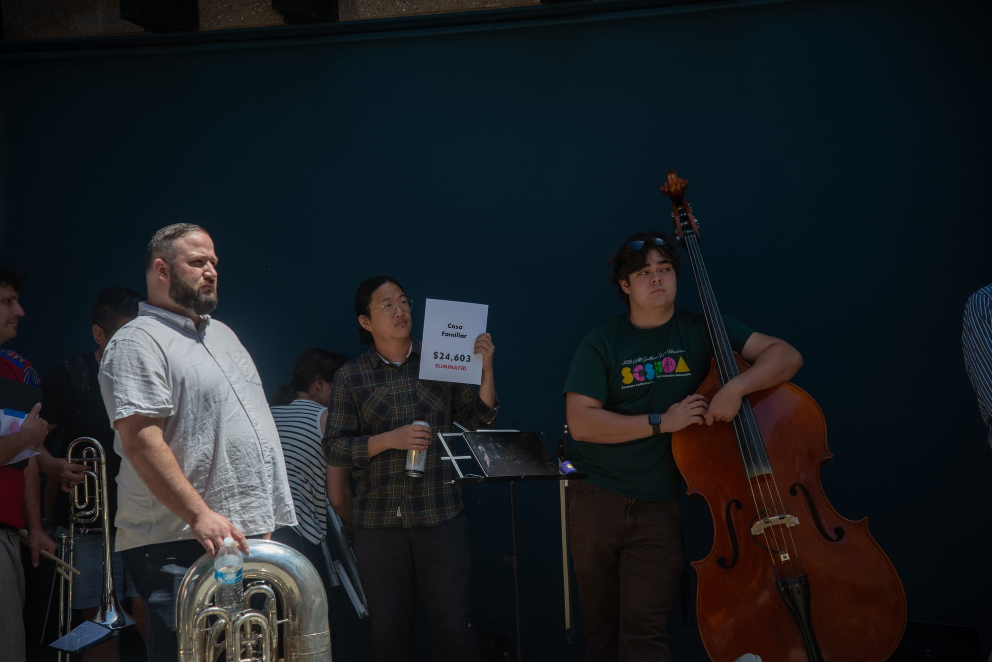 A man with an upright bass stands next to a man holding up a piece of paper that says Casa Familiar $24,603 eliminated. Another man with a tuba stands next to them.