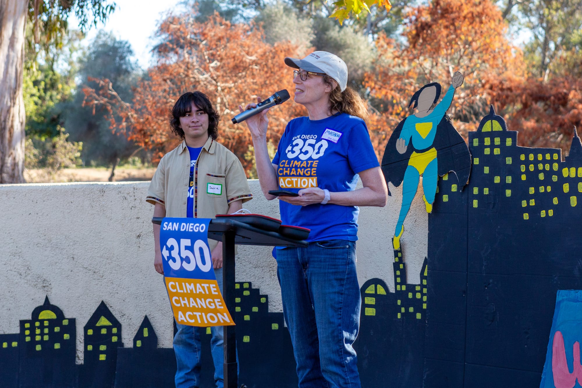 A woman in a blue shirt, blue jeans and a hat speaks through a microphone in front of a podium outside. 