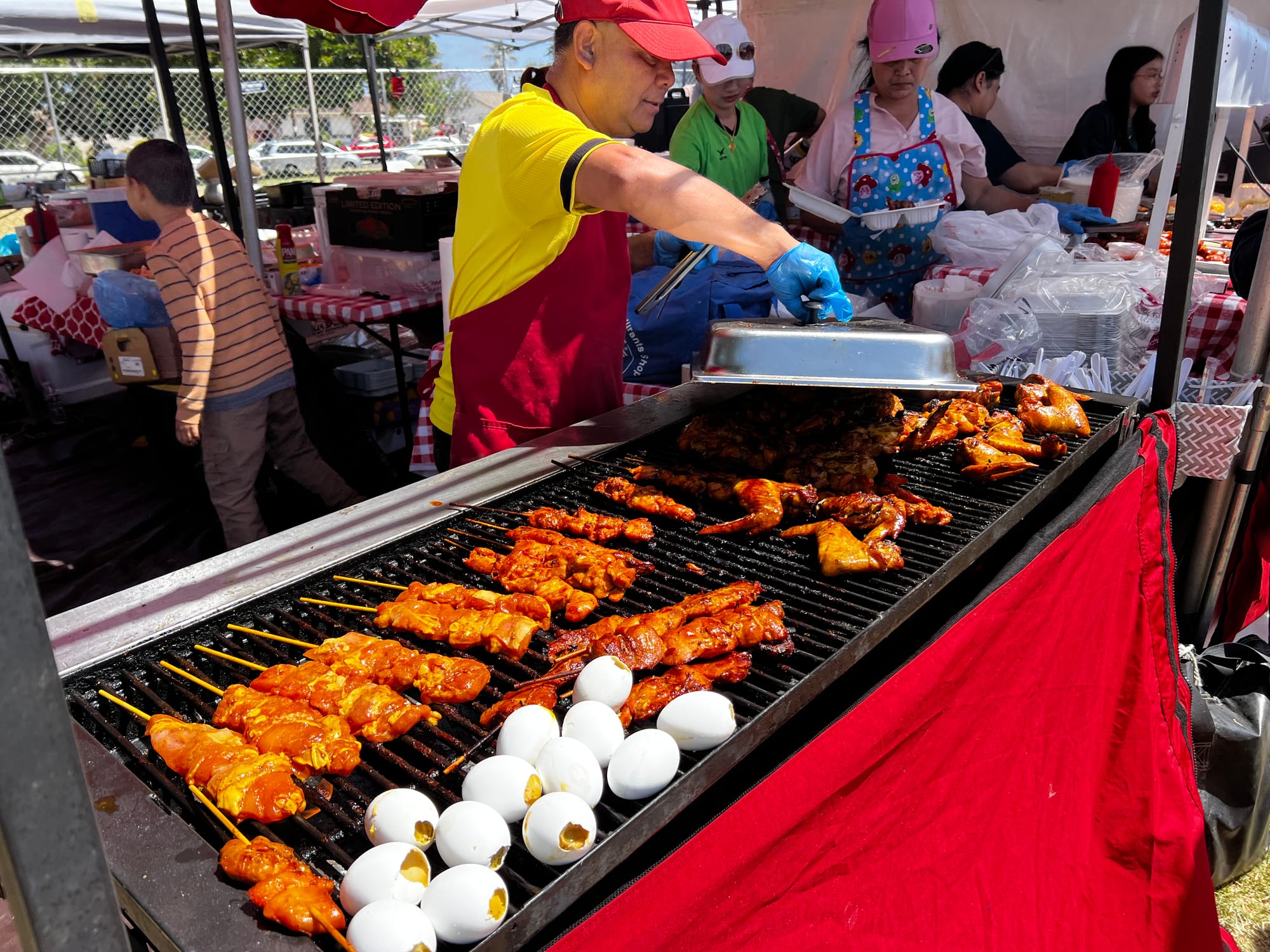 A person lifts a cover over food on a grill at an outdoor food event