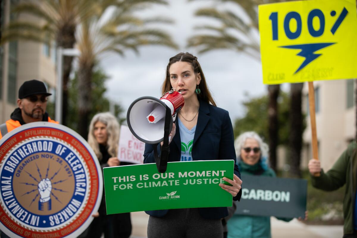 A woman in a dark blue blazer, standing in a crowd of demonstrators, speaks into a megaphone and holds a sign that reads, "This is our moment for climate justice".