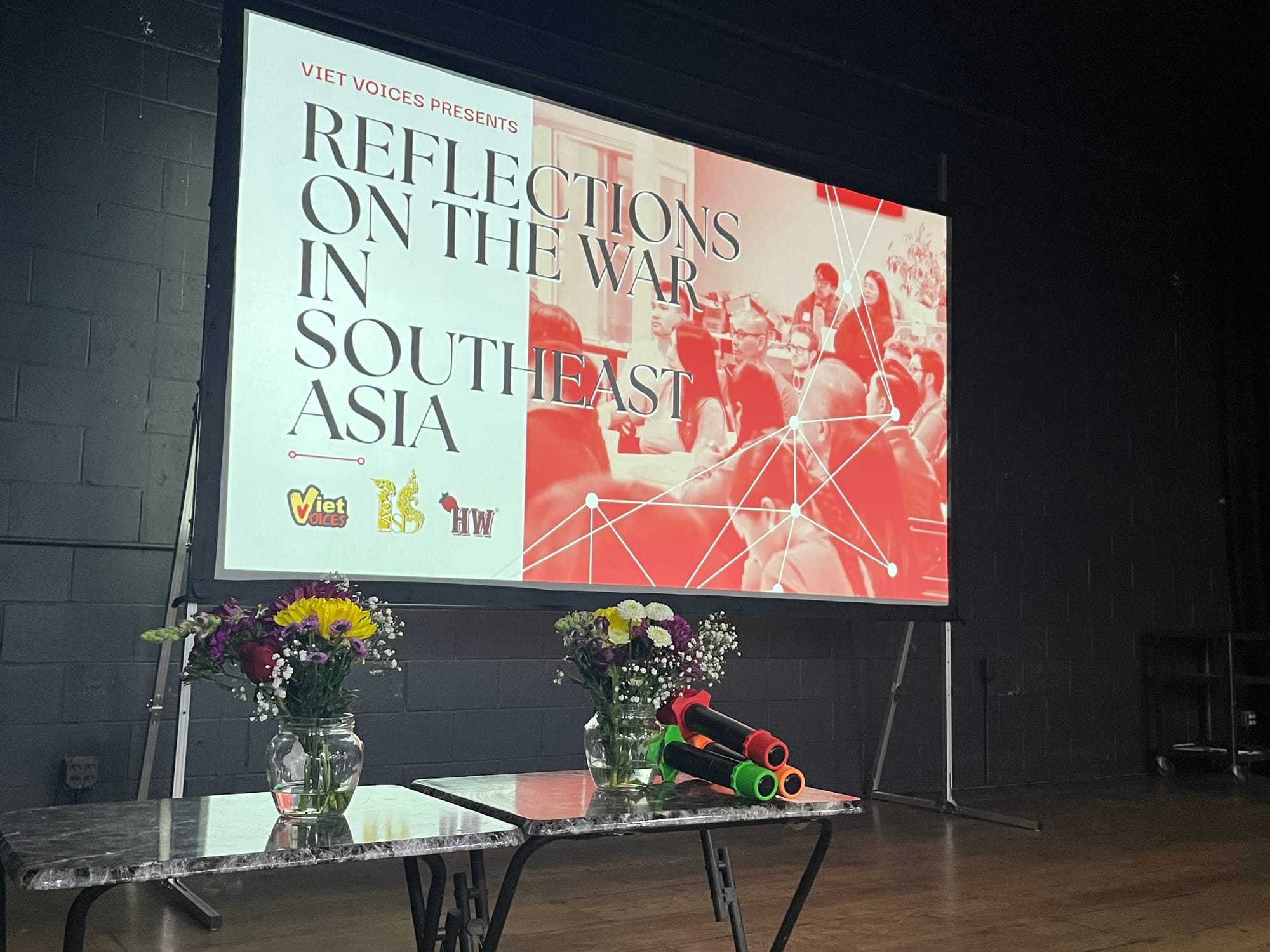 Two tables with assorted colorful flowers and microphones sit in front of a screen that reads "Reflections on the war in Southeast Asia"