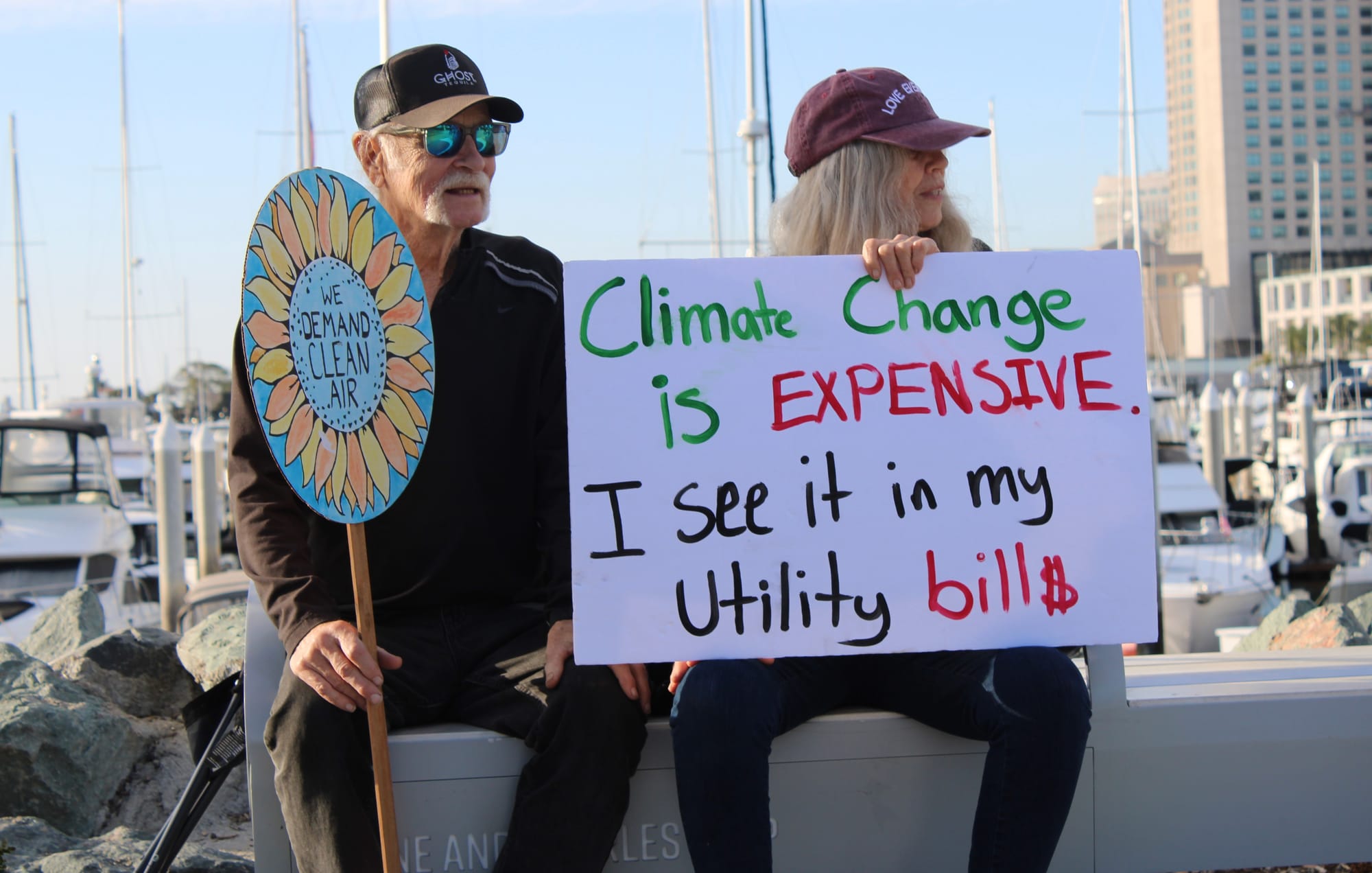 A man with a hat, sunglasses and beard holds a sign reading, "We Demand Clean Air," beside a woman in a hat holding a sign reading, "Climate change is Expensive. I see it in my utility bills."