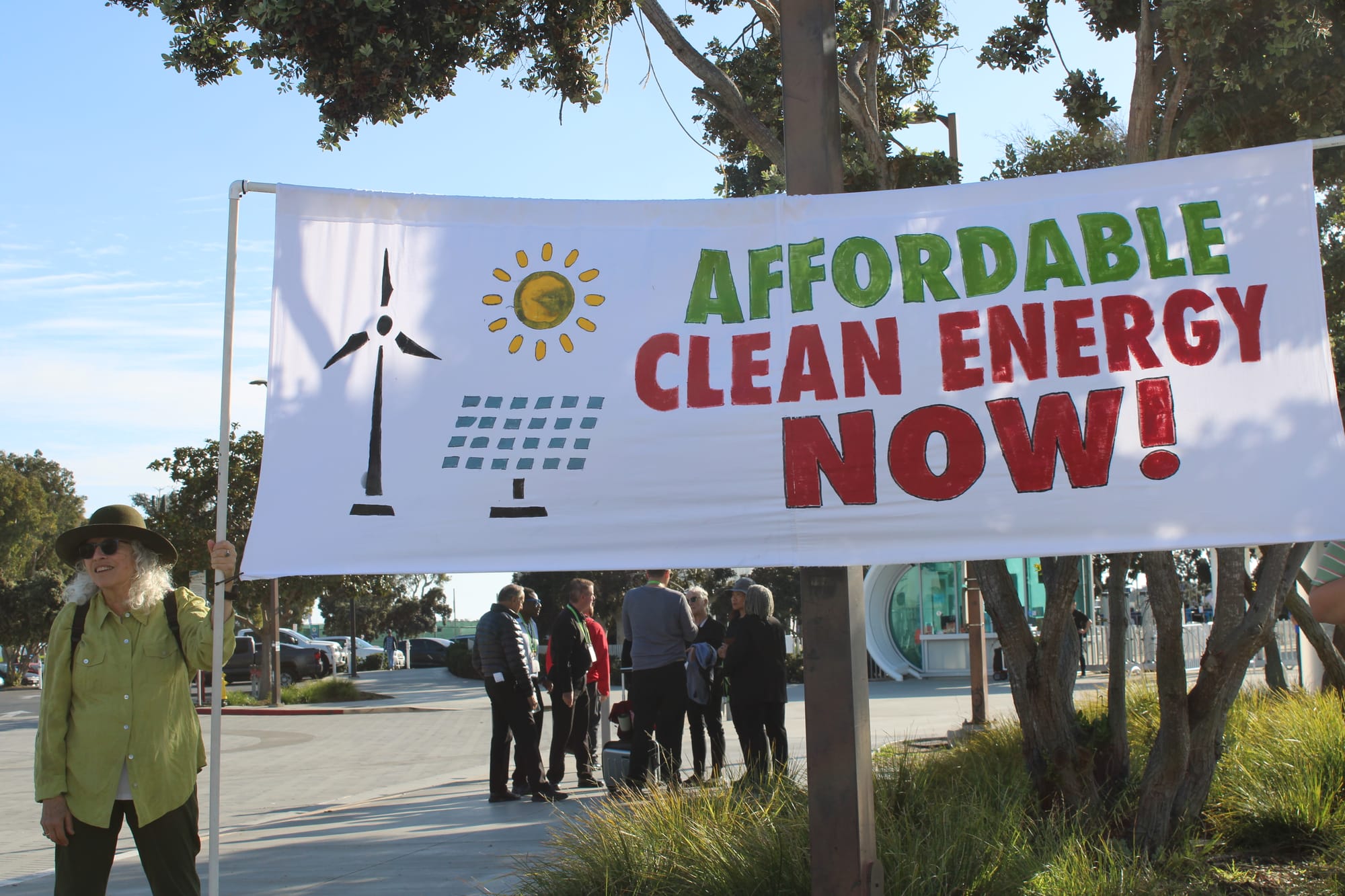 A woman in a green shirt and brown hat helps hold up one side of a large banner reading, "Affordable Clean Energy Now!"
