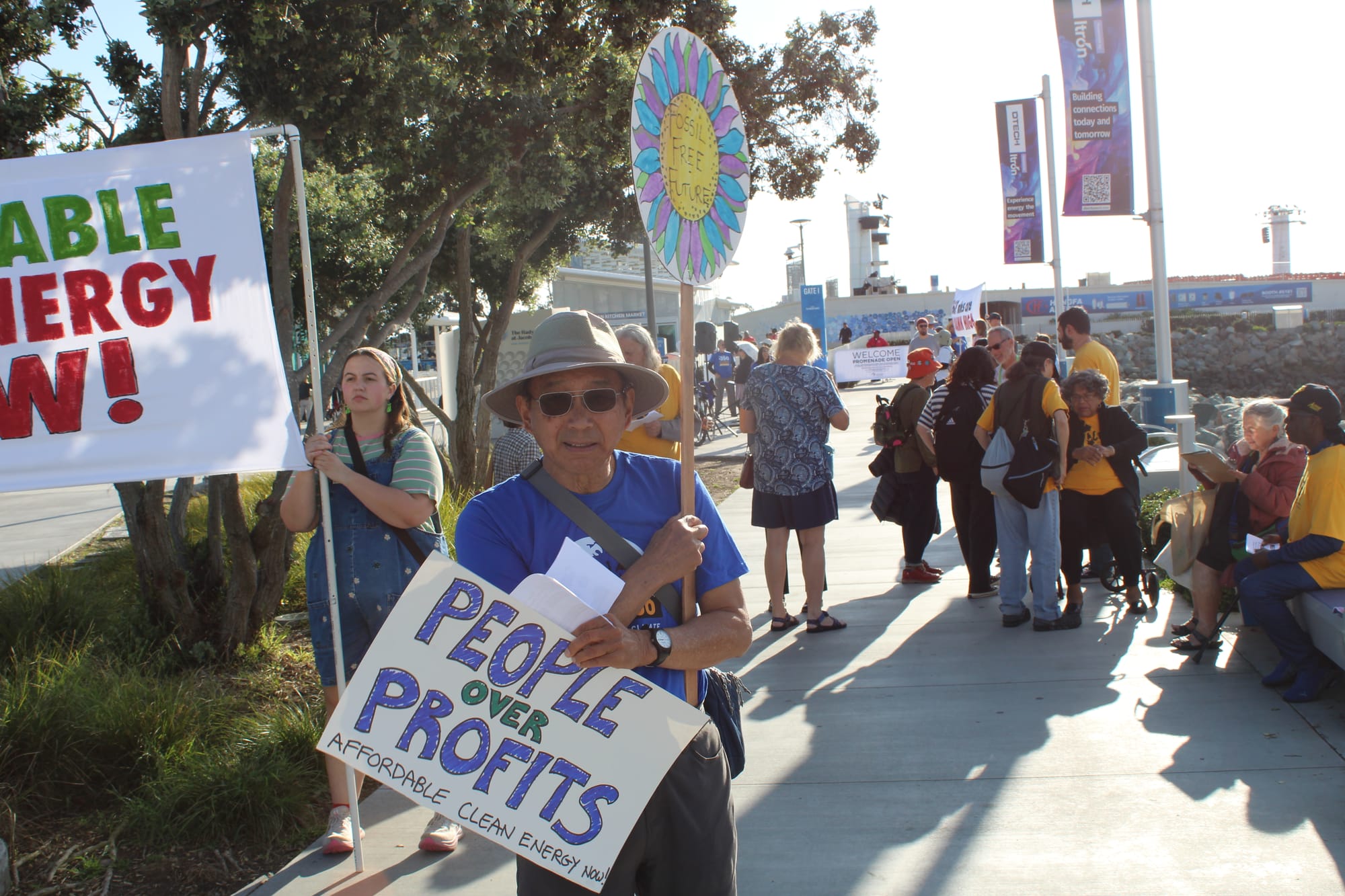 A man with a blue shirt and tan hat holds signs reading, "Fossil Free Future," and "People over profits."