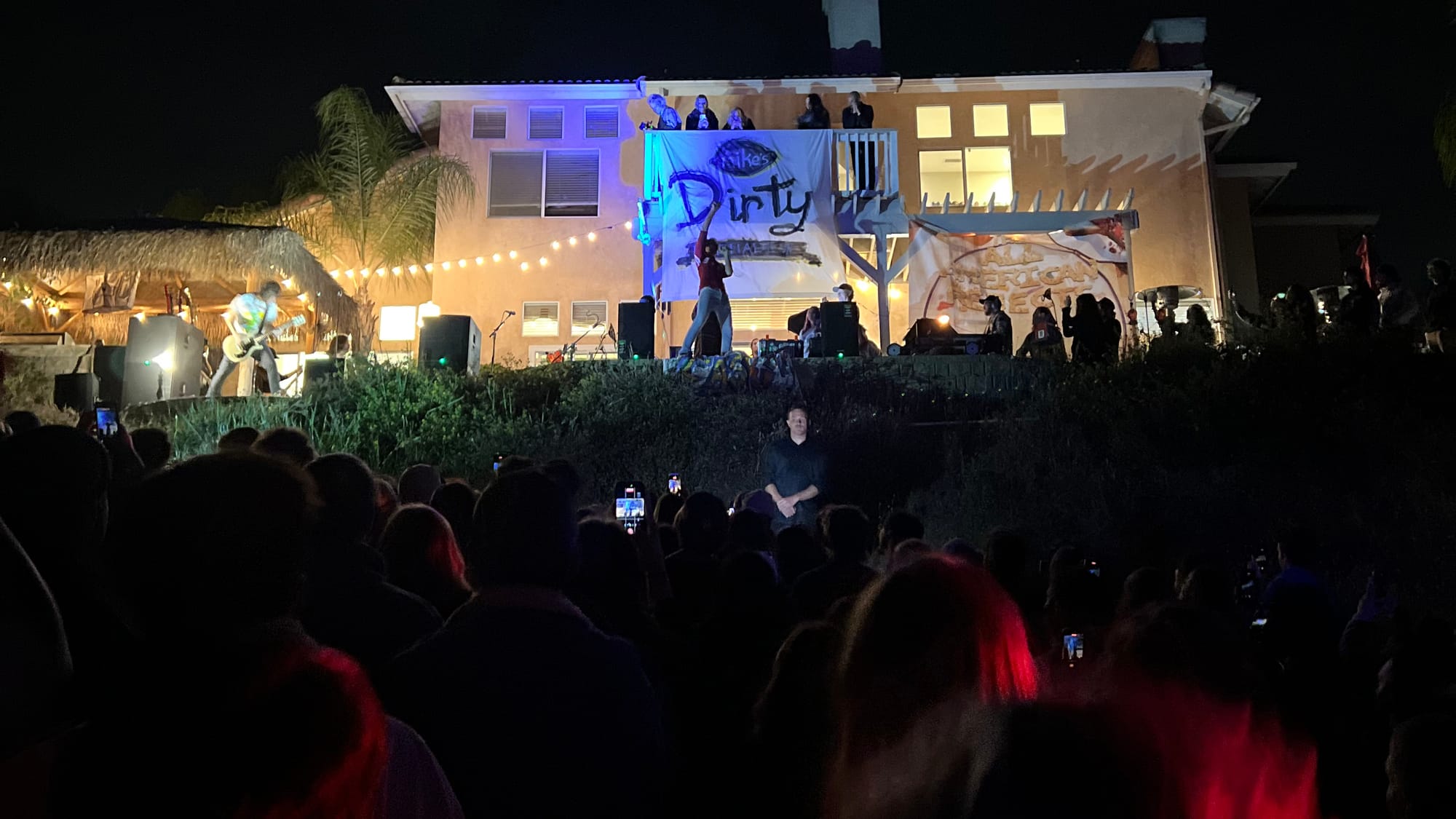 A band plays on a patio in front of a big crowd on a hill in the back of a house.