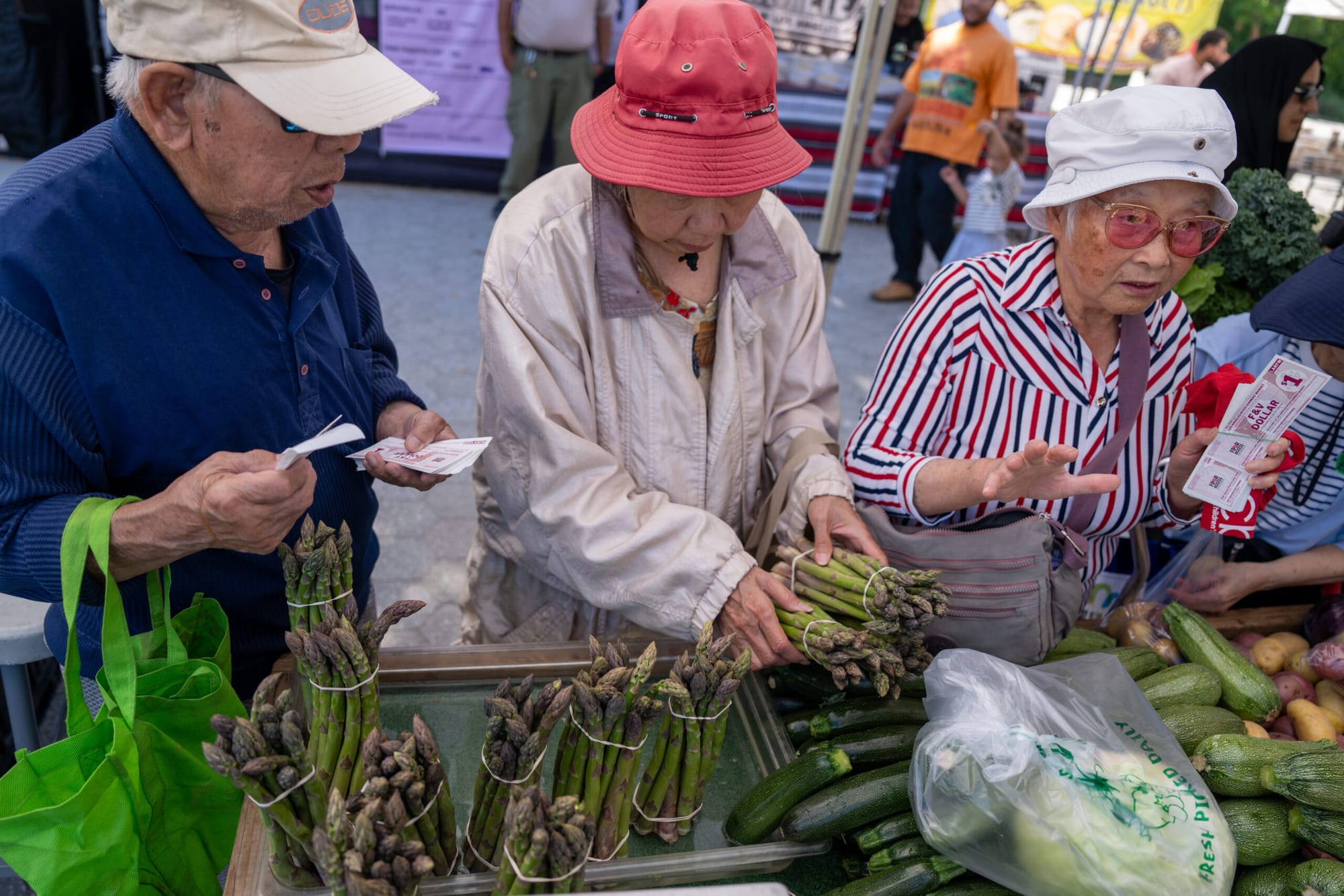 Three people hold asparagus and look at vegetables at a farmers market stand