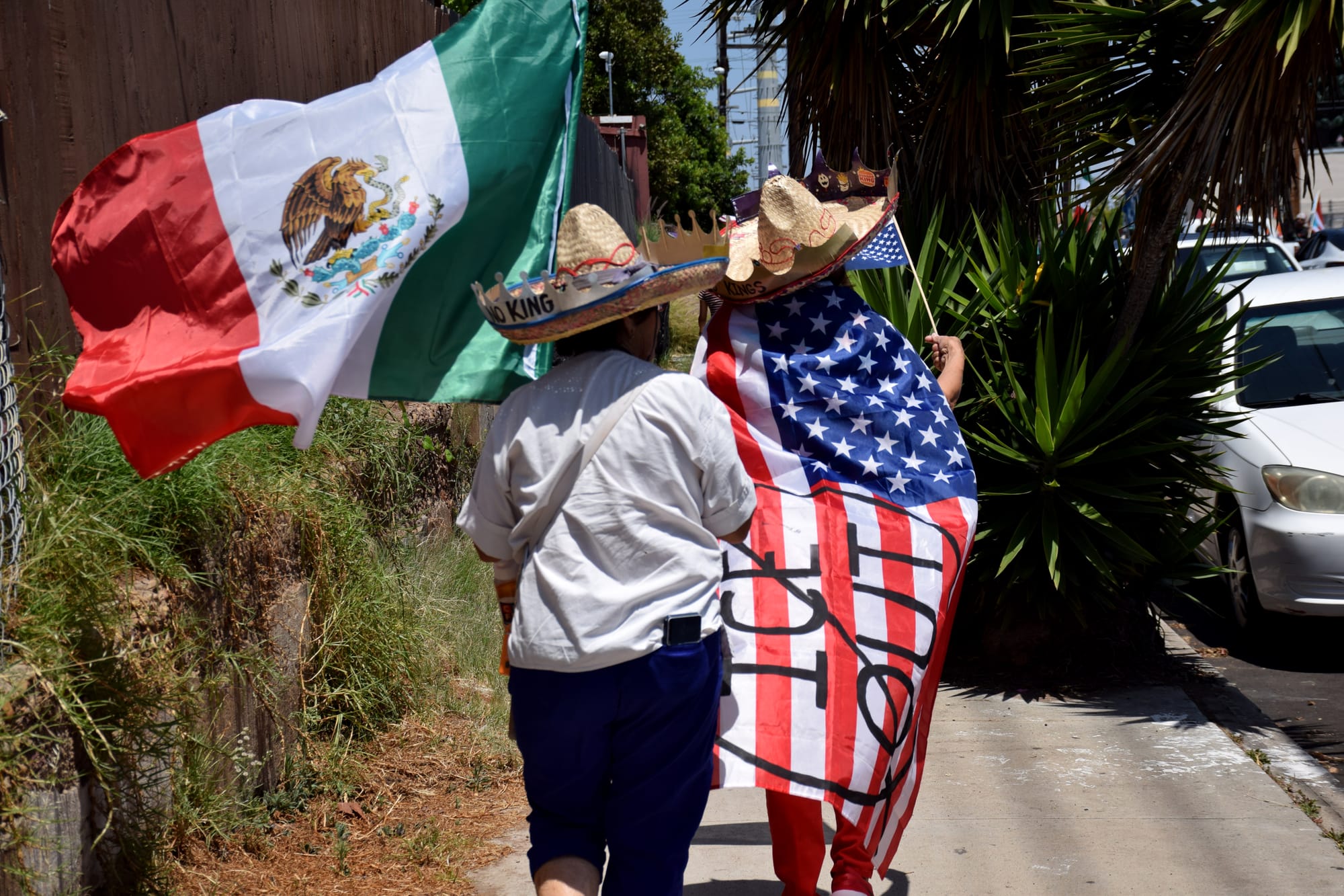 Two women wearing sombreros walk down the sidewalk. One carries a Mexican flag while the other wears an American flag as a cape that reads "ICE Out."