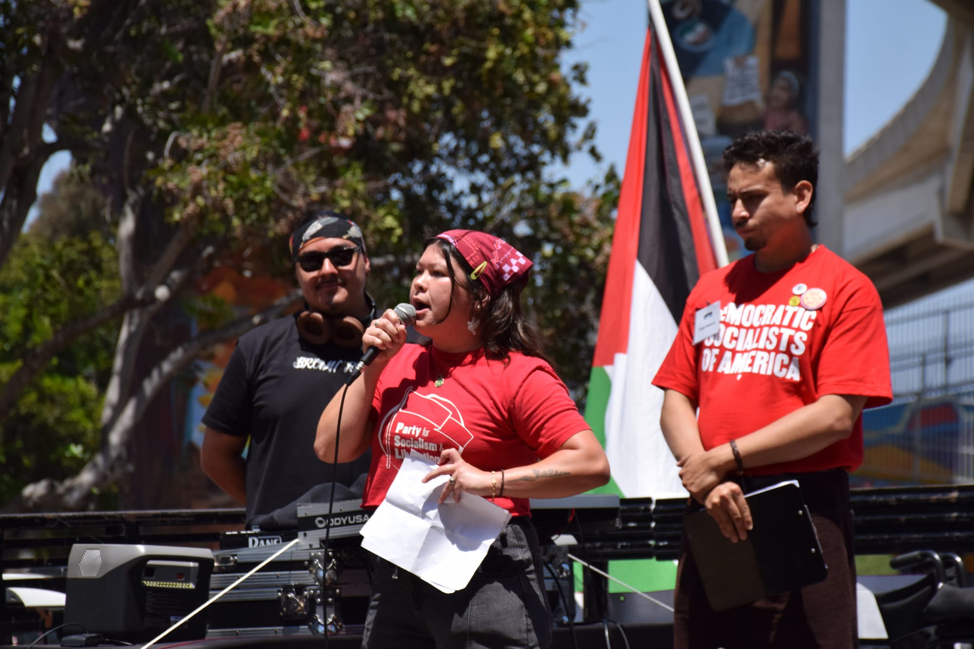 A person in a red t-shirt that reads "Party for Socialism & Liberation" speaks on a stage during a protest