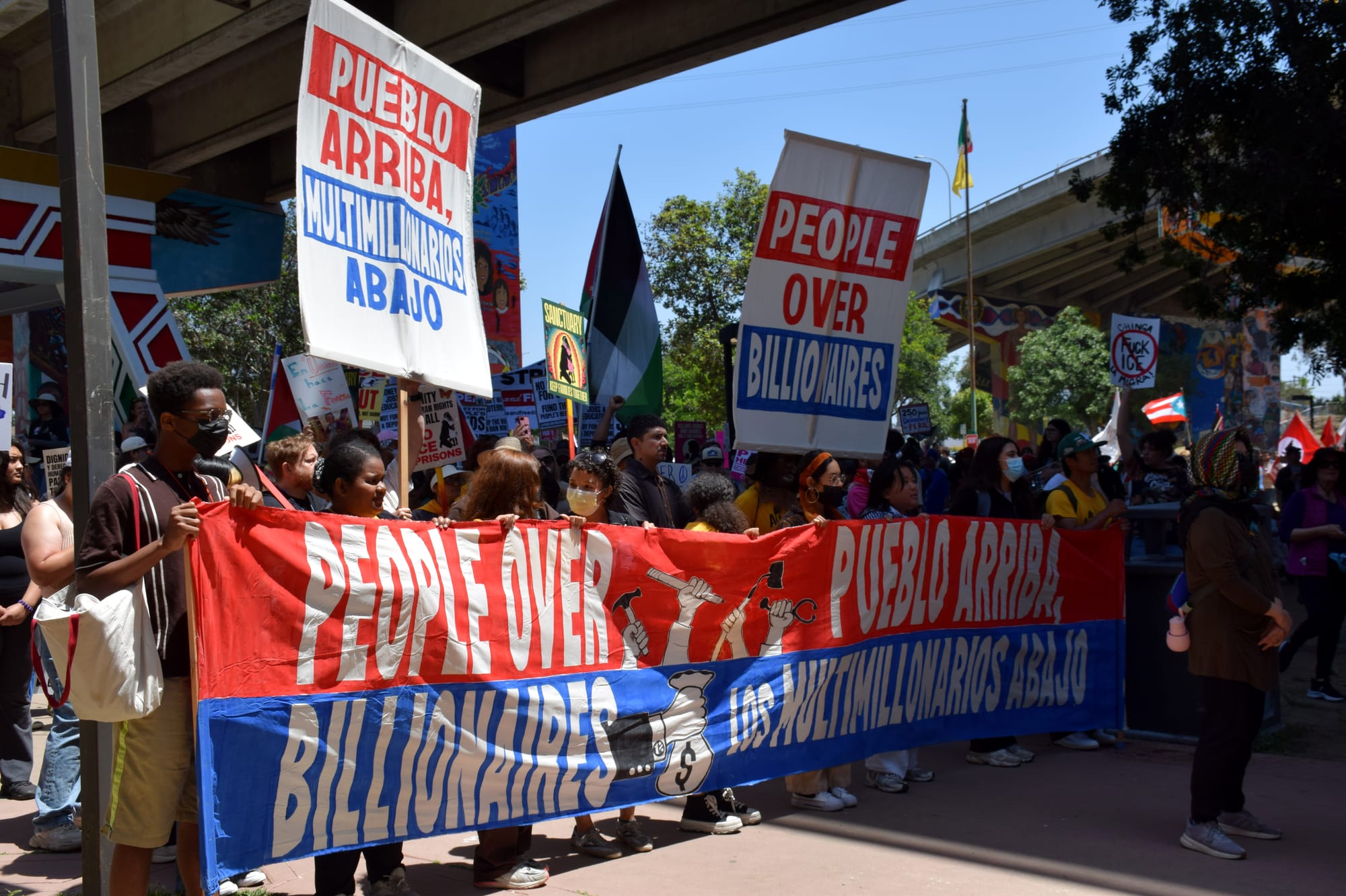 A group of protestors in hold a red, white and blue banner and signs that read “People over billionaires” and “Pueblo arriba, los multimillonarios abajo (The people on top, the billionaires on the bottom.”