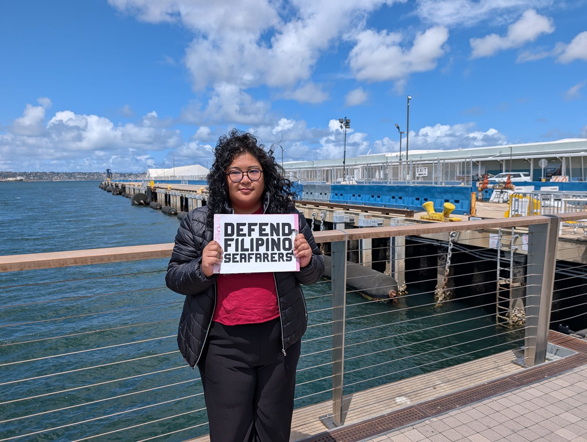 A woman in a red shirt and black jacket holds a sign that says Defend Filipino Seafarers
