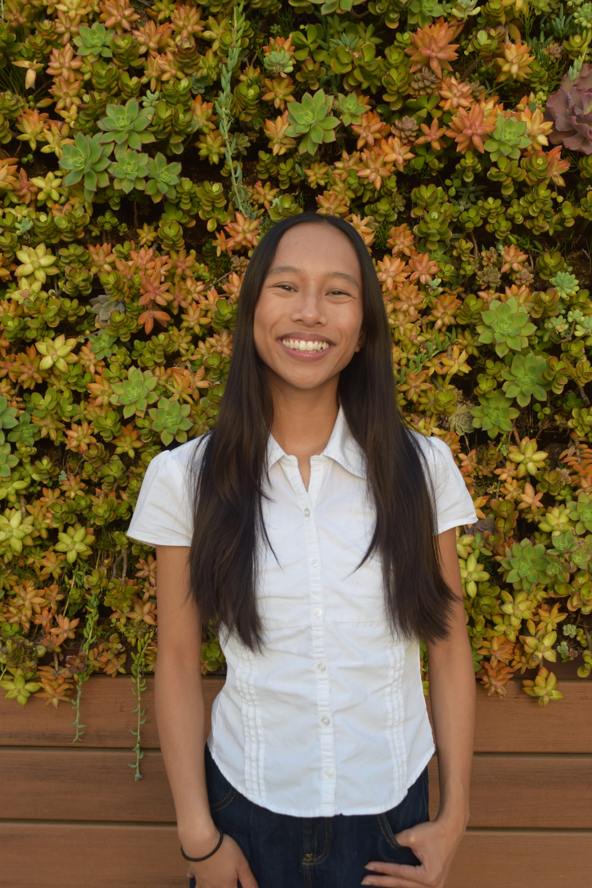 Picture of someone smiling, wearing a white button in front of a succulent plant wall