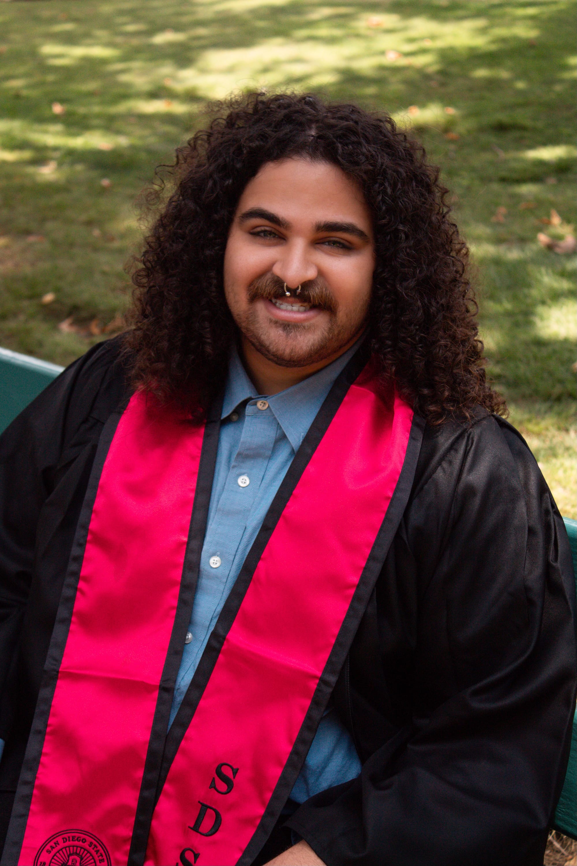 Picture of someone with curly hair wearing a black graduation gown and a red stole that reads "San Diego State University" and "SDSU"