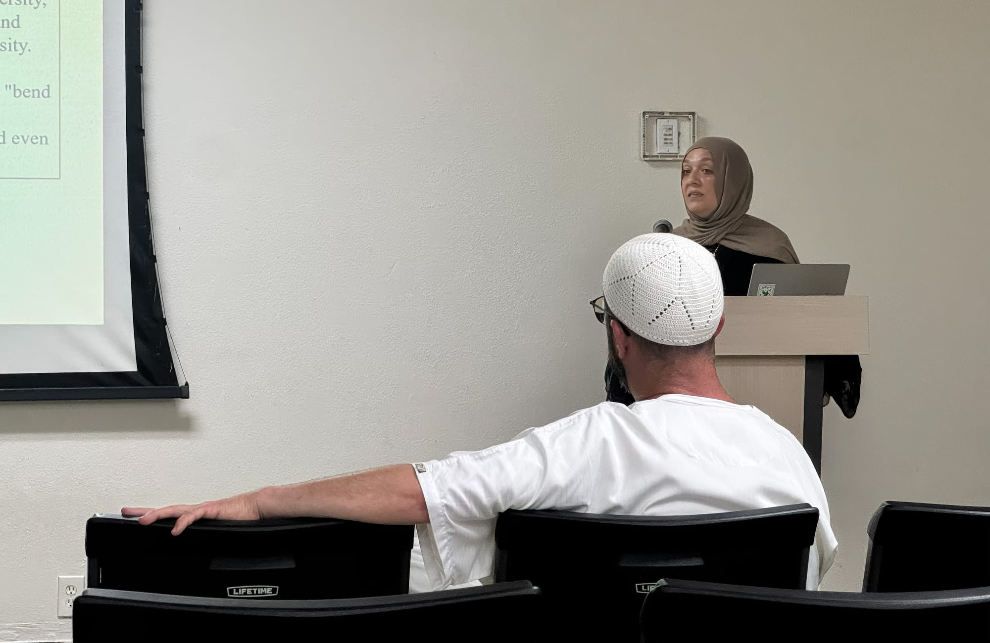 A woman in a beige hijab and black clothing stands at a podium while a man wearing white sits in front of her in the audience