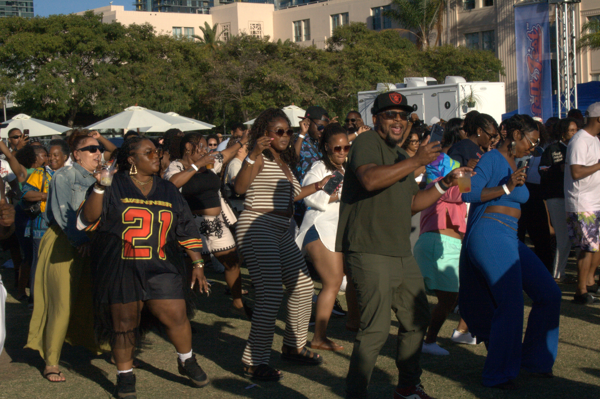 People line dance at an outdoor music festival in front of trees and a building