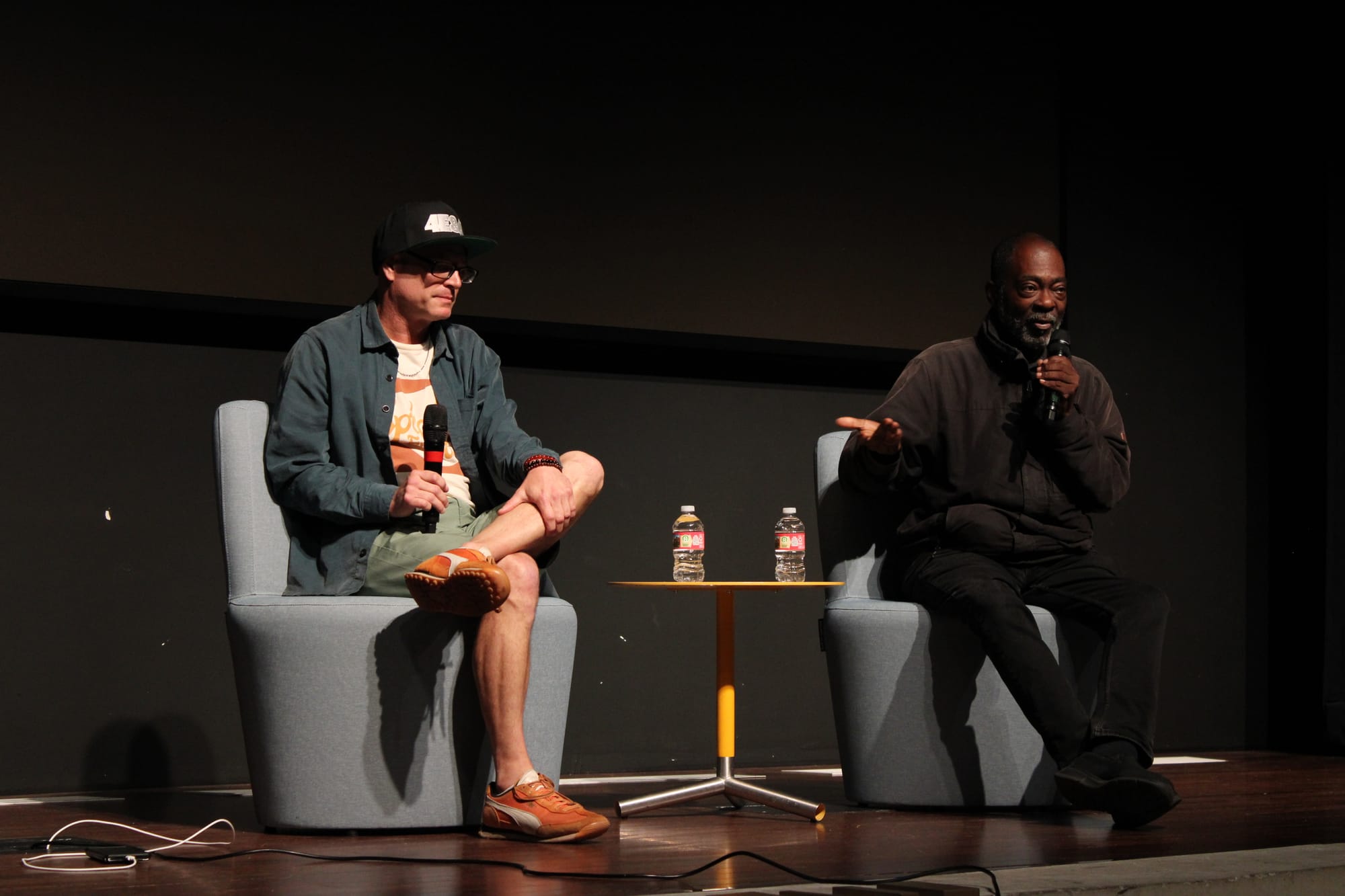 Two men sit in gray chairs on stage holding microphones.