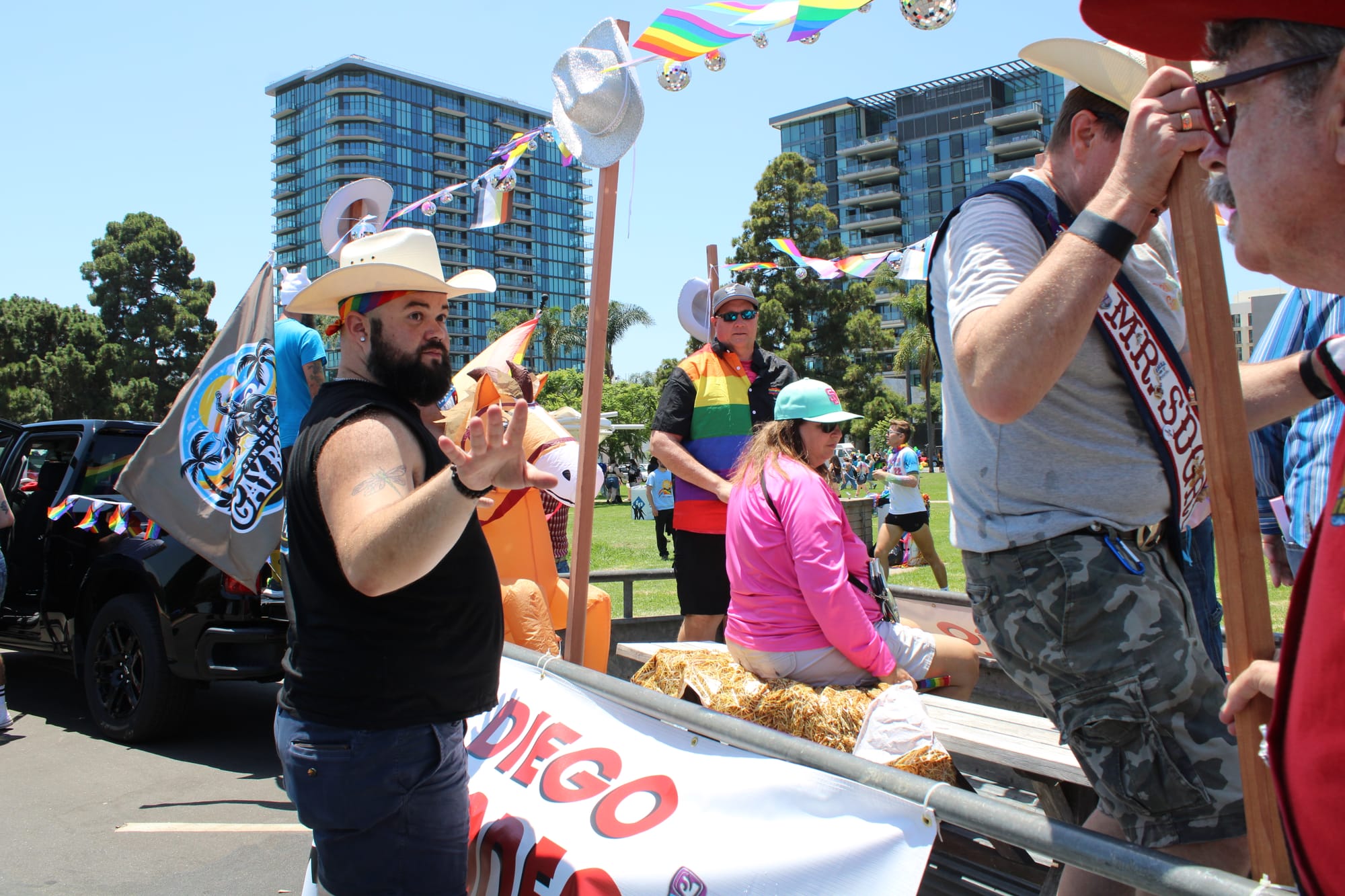 A person in a cowboy hat and rainbow bandana stands in front of a trailer with people on it