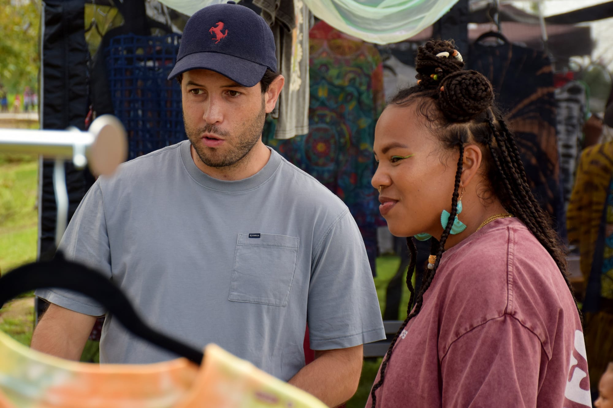 Two people talk at an outdoor vendor booth with grass and clothes in the background
