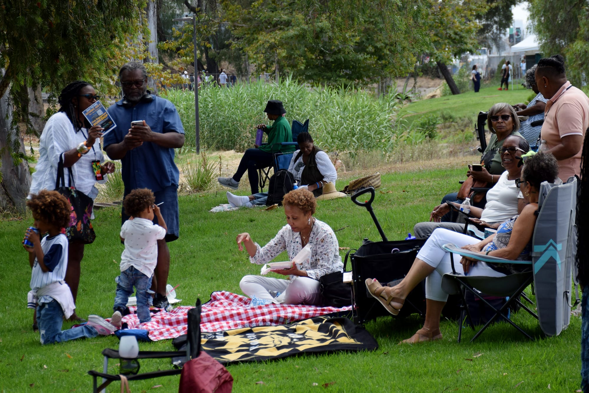 A group of people sit and stand around picnic blankets and folding camping chairs in a grassy field