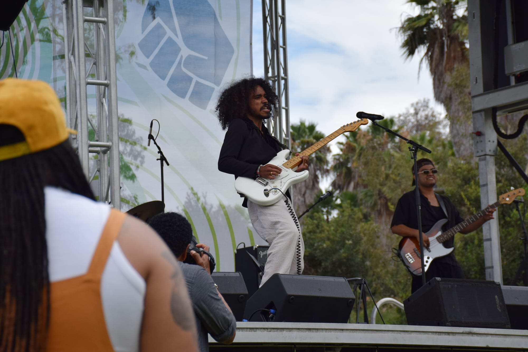 One person plays a bass guitar and another plays the guitar on stage in front of trees at an outside music festival