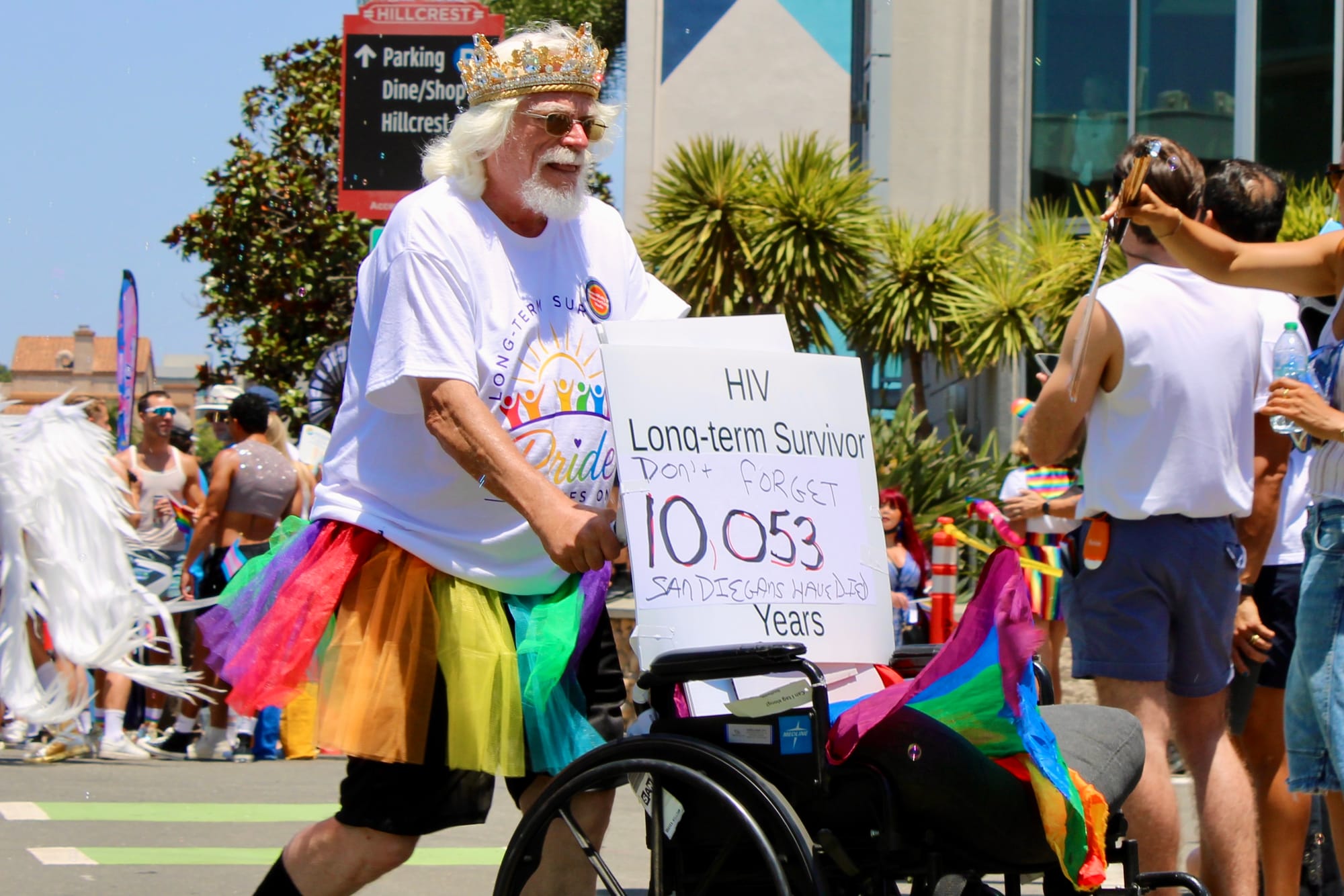 A person with a gray beard and a rainbow tutu pushes a wheelchair with a sign that says, "HIV Long-term survivor, DOn't gorget 10,053 San Diegans have died"