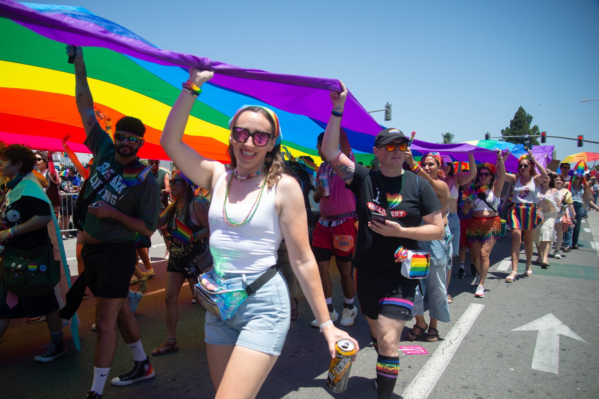 A person in a white shrt and jean shorts stands in the sunshine holding up the purple end of the Pride flag
