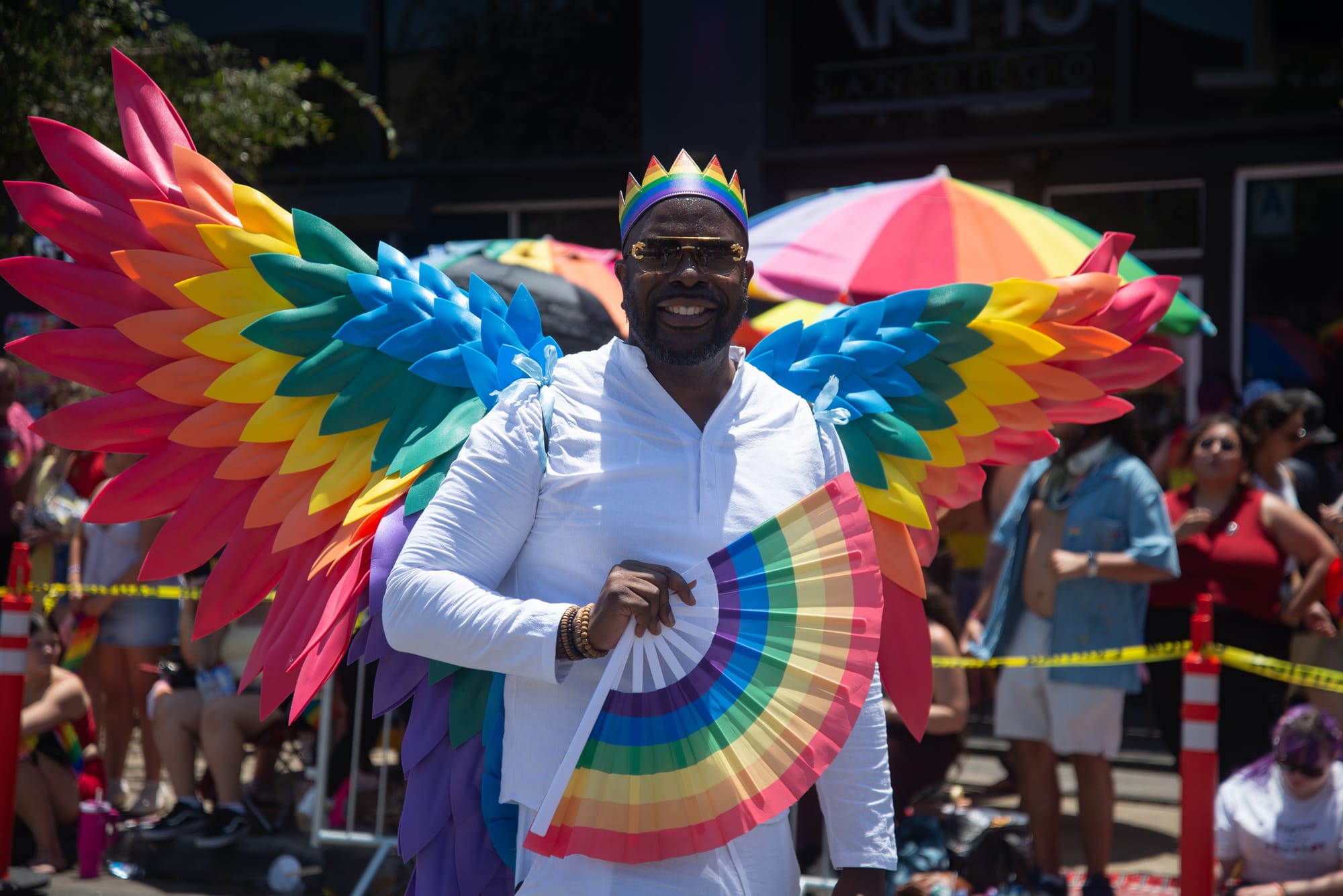 A man in white clothing wears rainbow wings and carries a rainbow fan