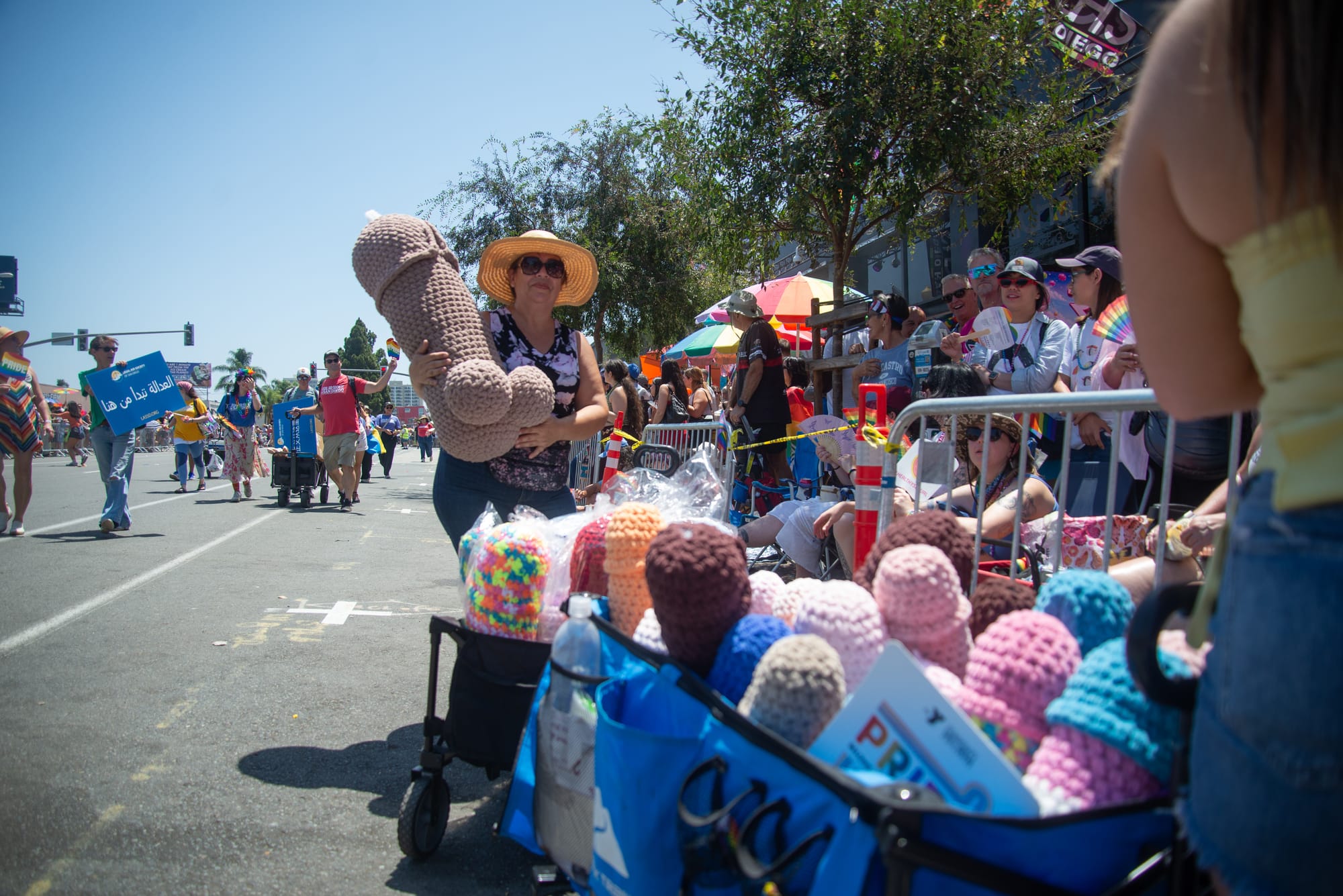 A woman in a hat holds up a large penis shaped pillow. In front of her are two wagons filled with penis pillows