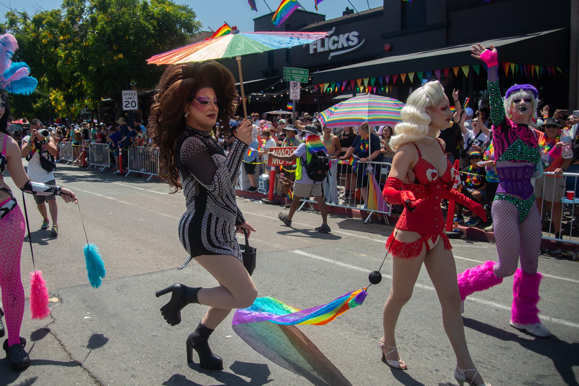A drag queen with large brown hair and a black dress and black boots holds on leg up while shading herself with a rainbow umbrella