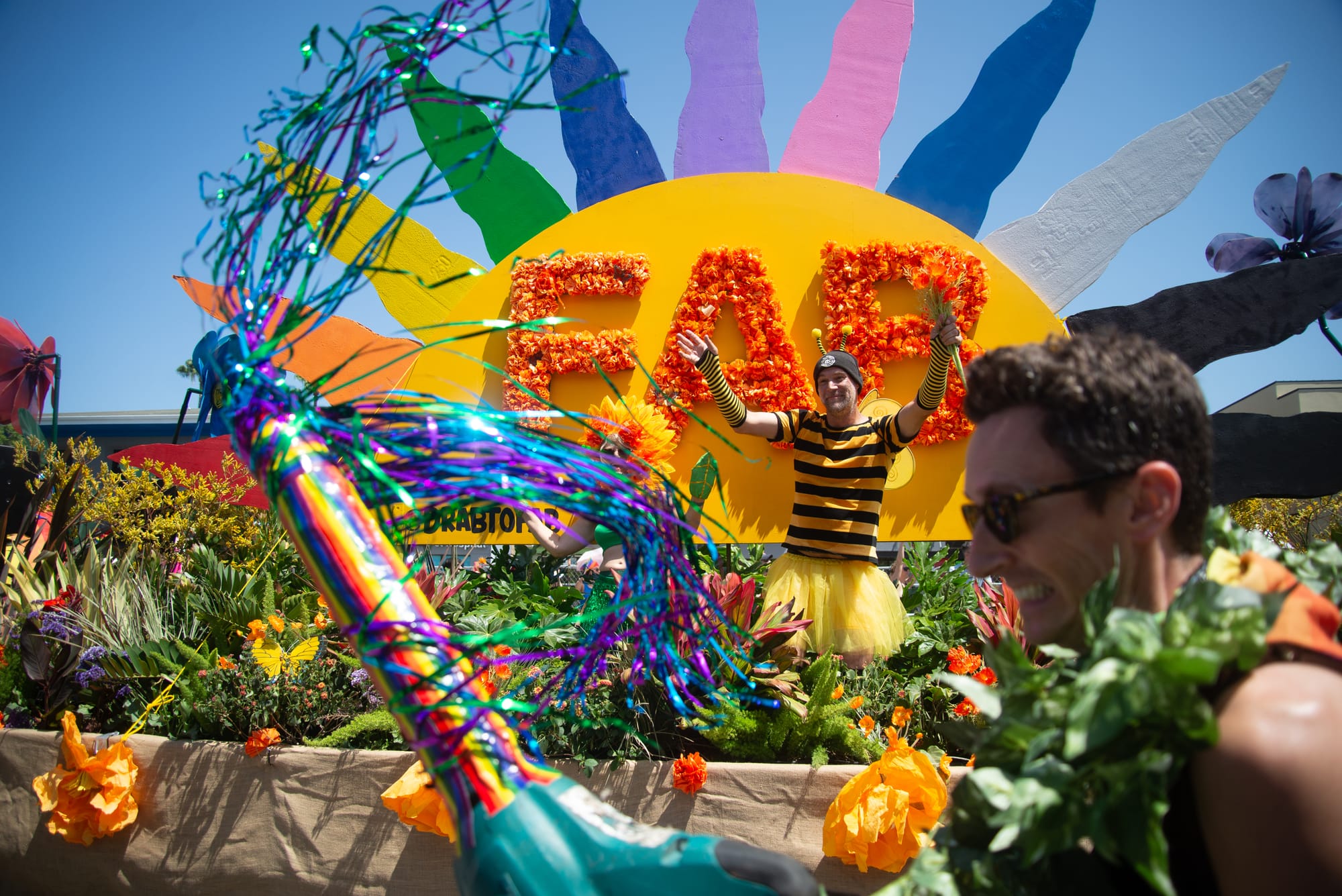A person wearing a bee costume waves from a float filled with plants and a sun that says FAB. In front, a person holds a decorated leaf blower.