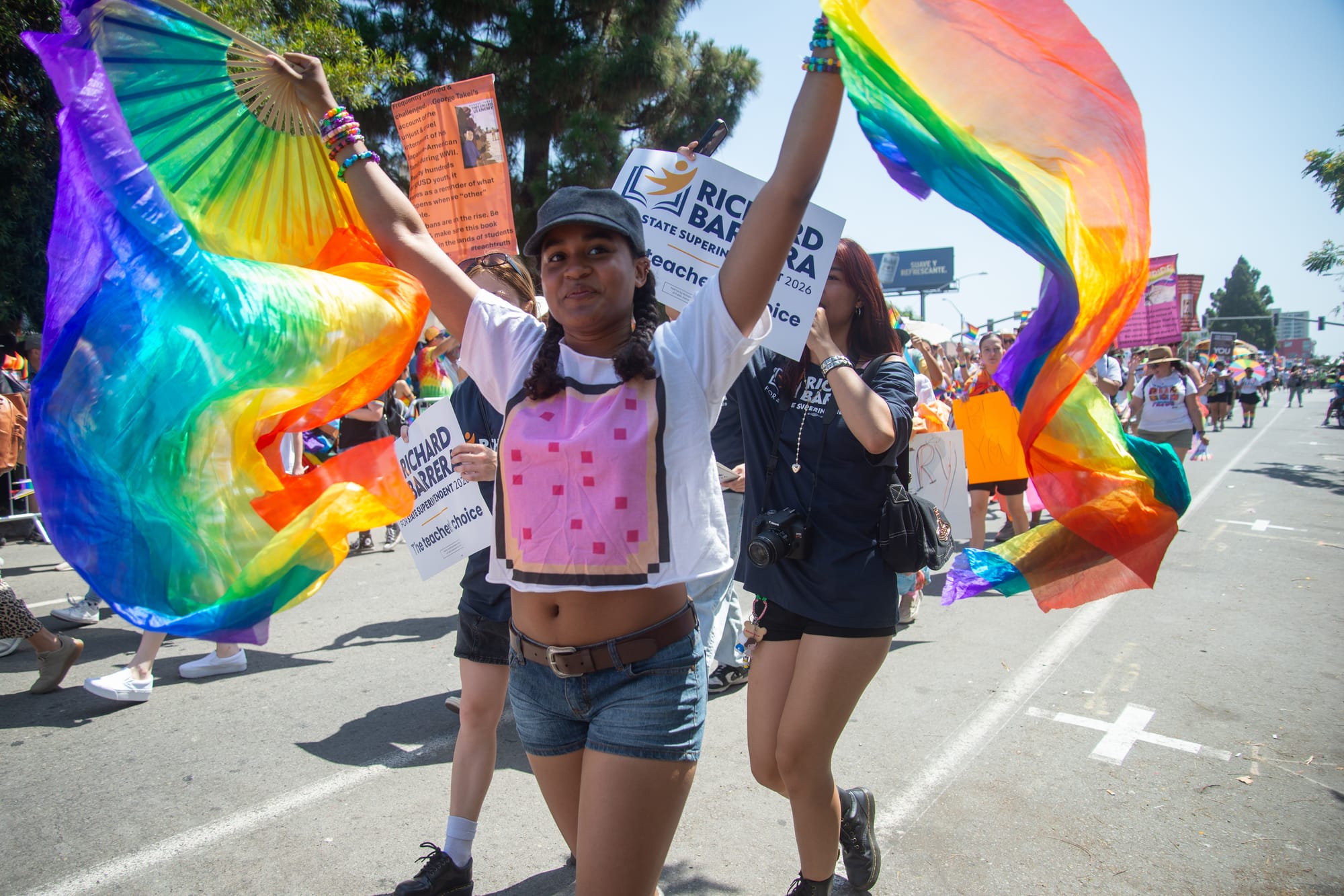 A person in a white shirt and jean shorts holds hands high with rainbow fans in each