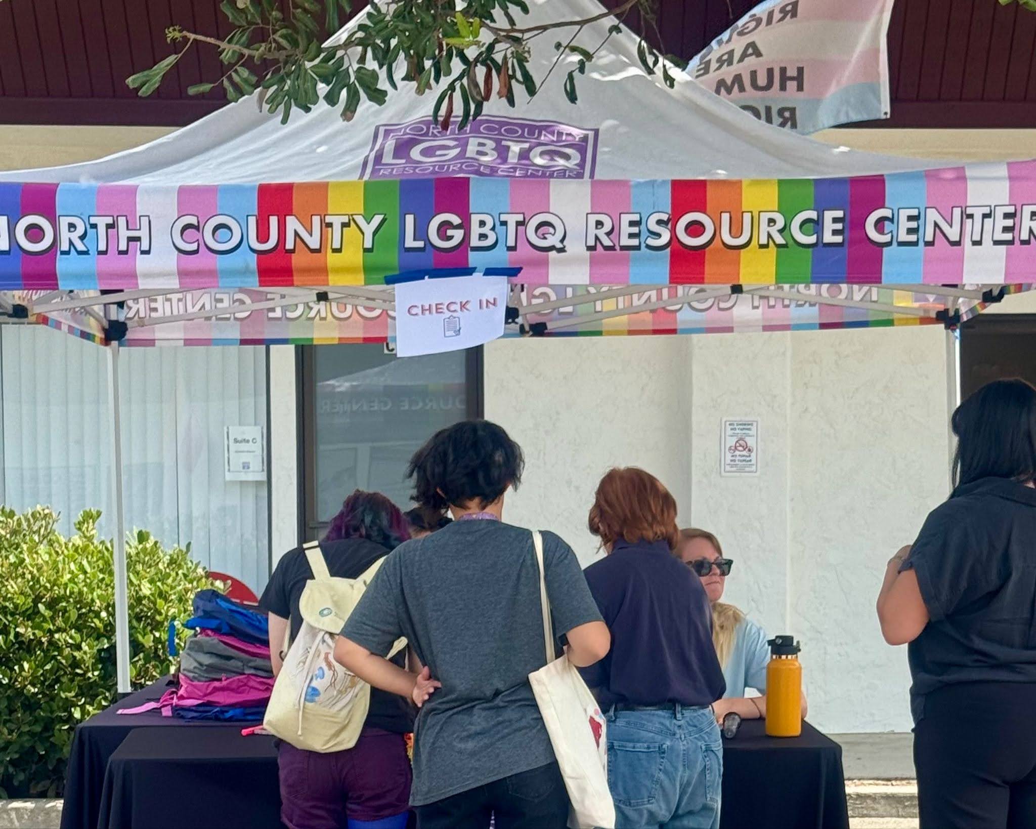 Three young people face away from the camera and toward a table with people sitting down on the other side. They are all under a tent that reads "North County LGBTQ Resource Center" with rainbow/pride color stripes.