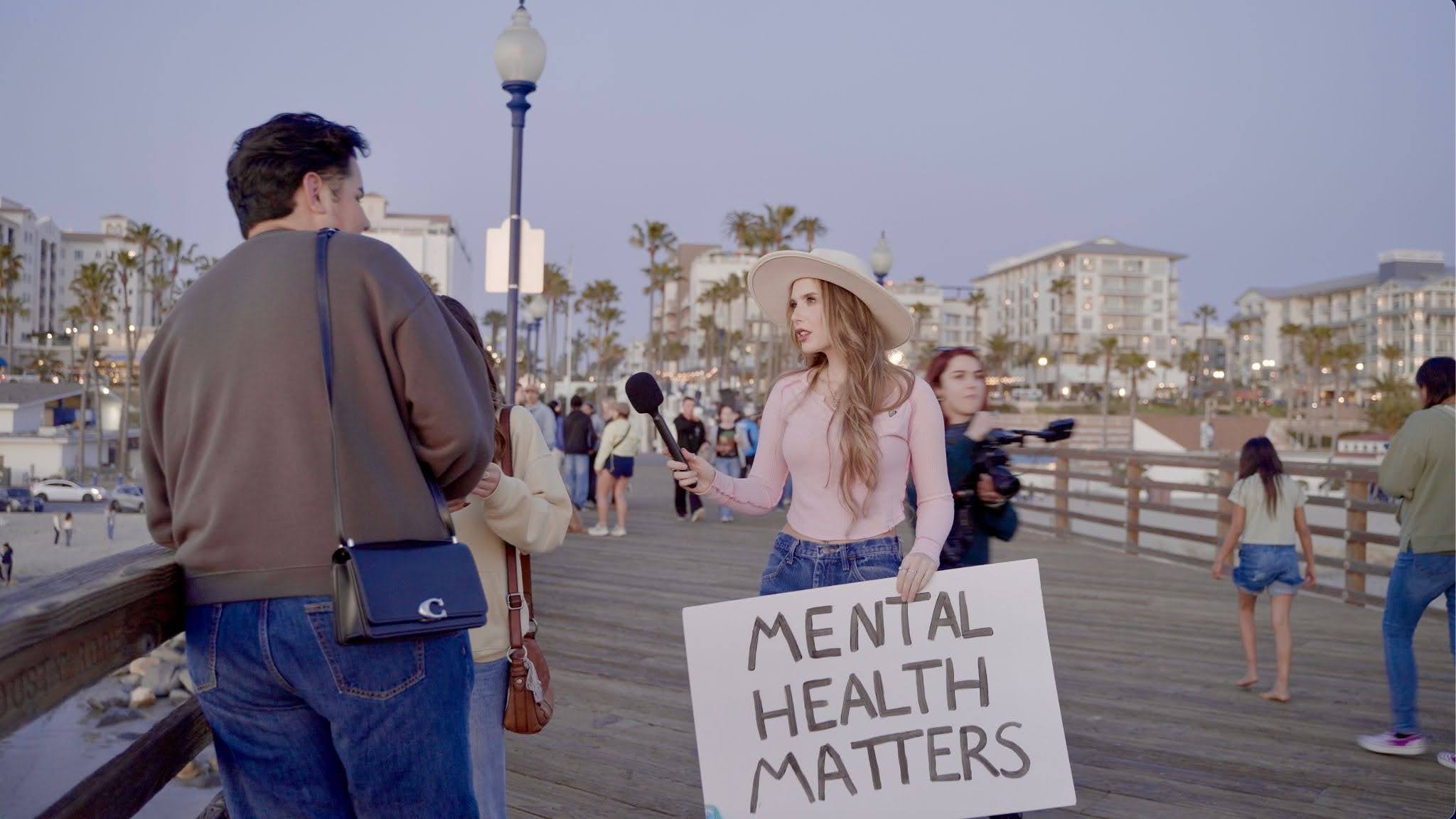 A woman holds a sign that reads "mental health matters" and a microphone up to a person on a busy boardwalk.