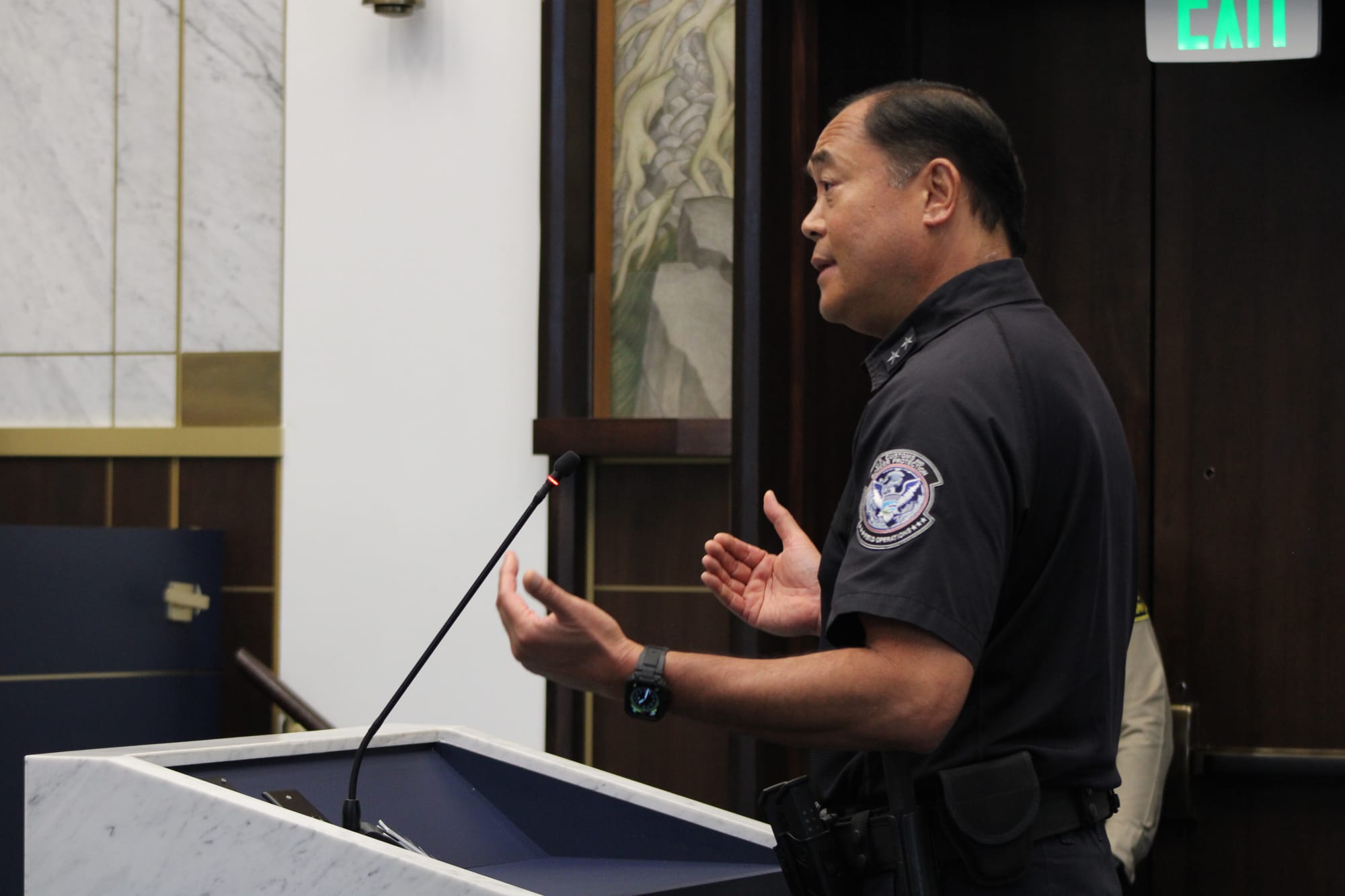 A man in a Customs and Border Protection uniform stands at a podium with his hands open