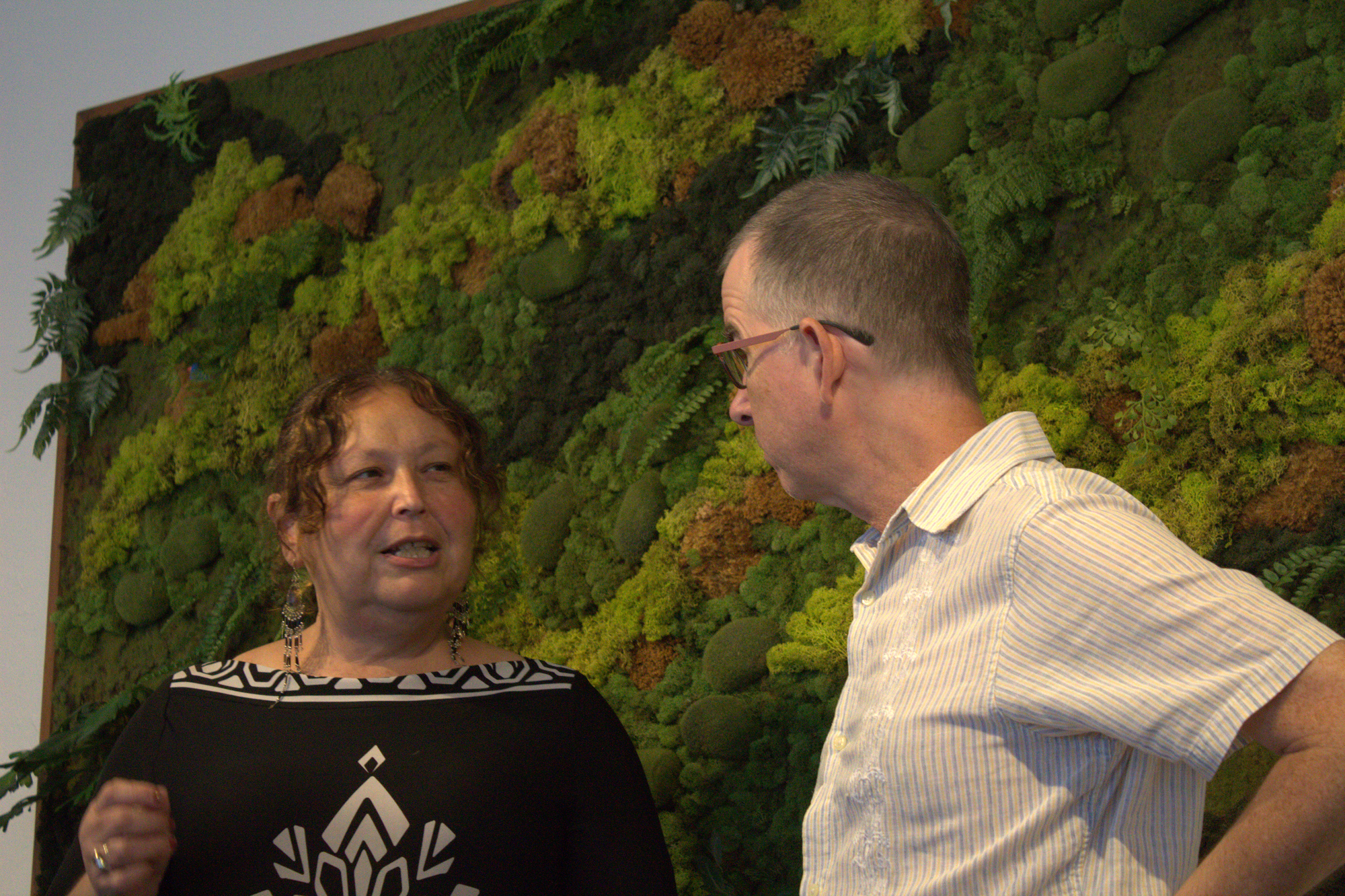 Two people talk in front of a textured moss art piece. 