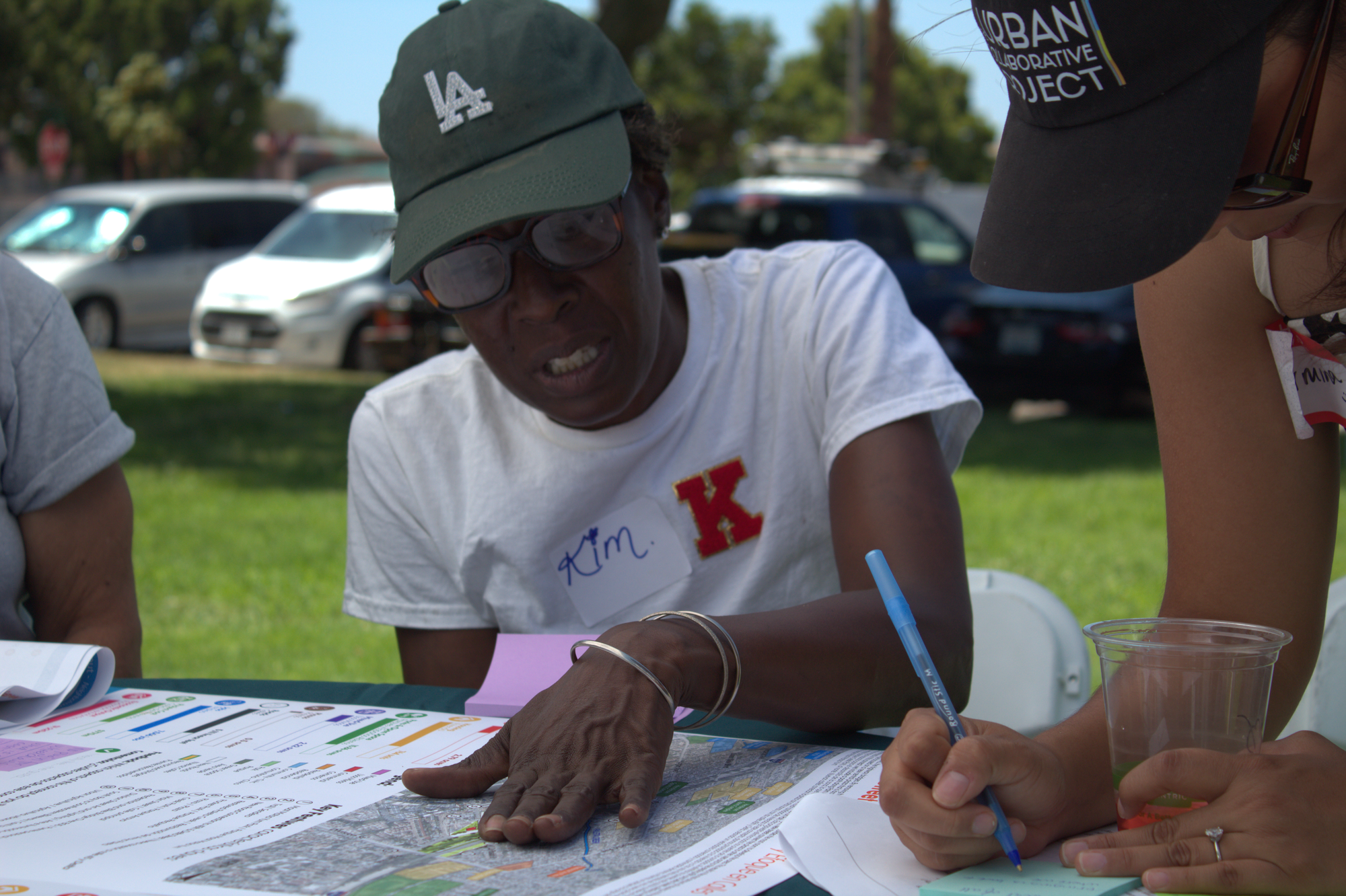 A person off to the side of the pictures is writing notes on a sticky note as a person talks while touching a map proposal. 