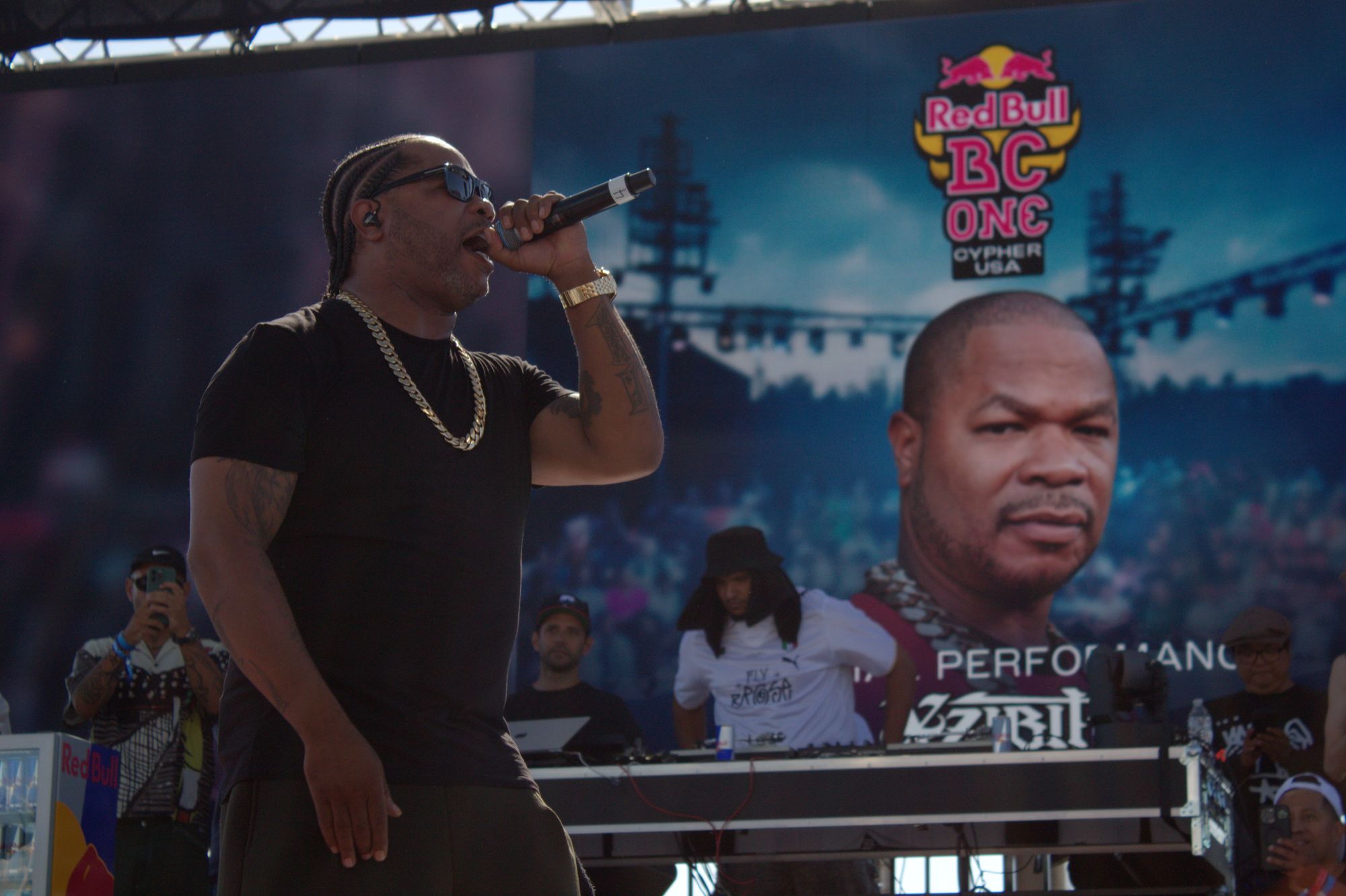 A person stands on stage in a black tee with gold chain holding a microphone