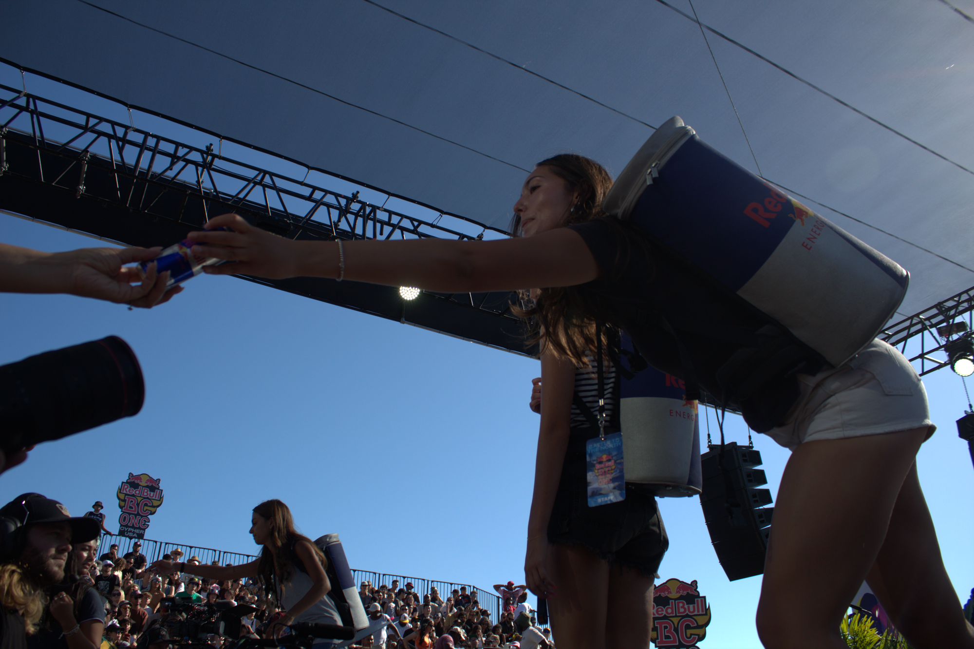 A girl with a Red Bull can-shaped backpack outstretches her arm to hand a Red Bull to someone grabbing it off to the side of the stage.