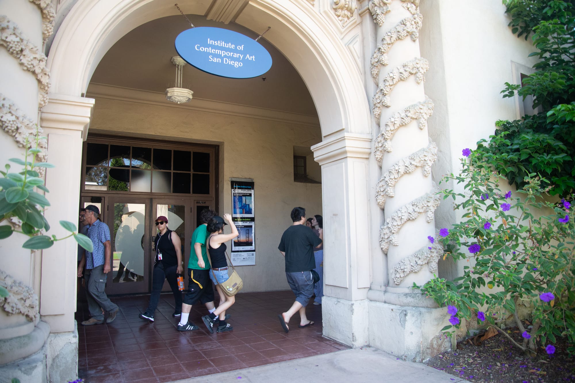 People walk in front of a glass door on a brown building and above them is a blue sign with the name of the museum