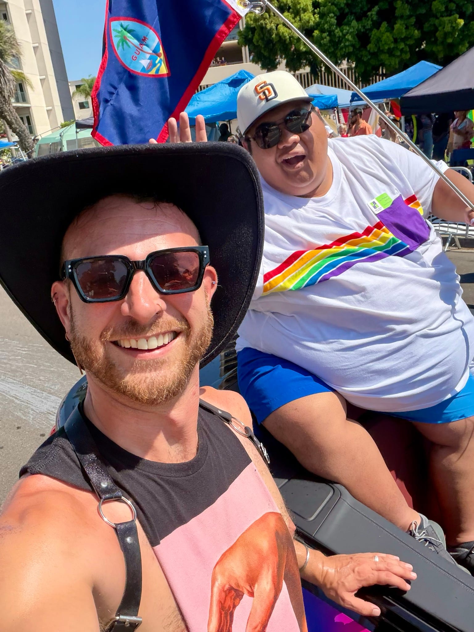 A man in a black hat and sleeveless shirt takes a selfie with a man in a white shirt with a rainbow across the front holding a Guam flag