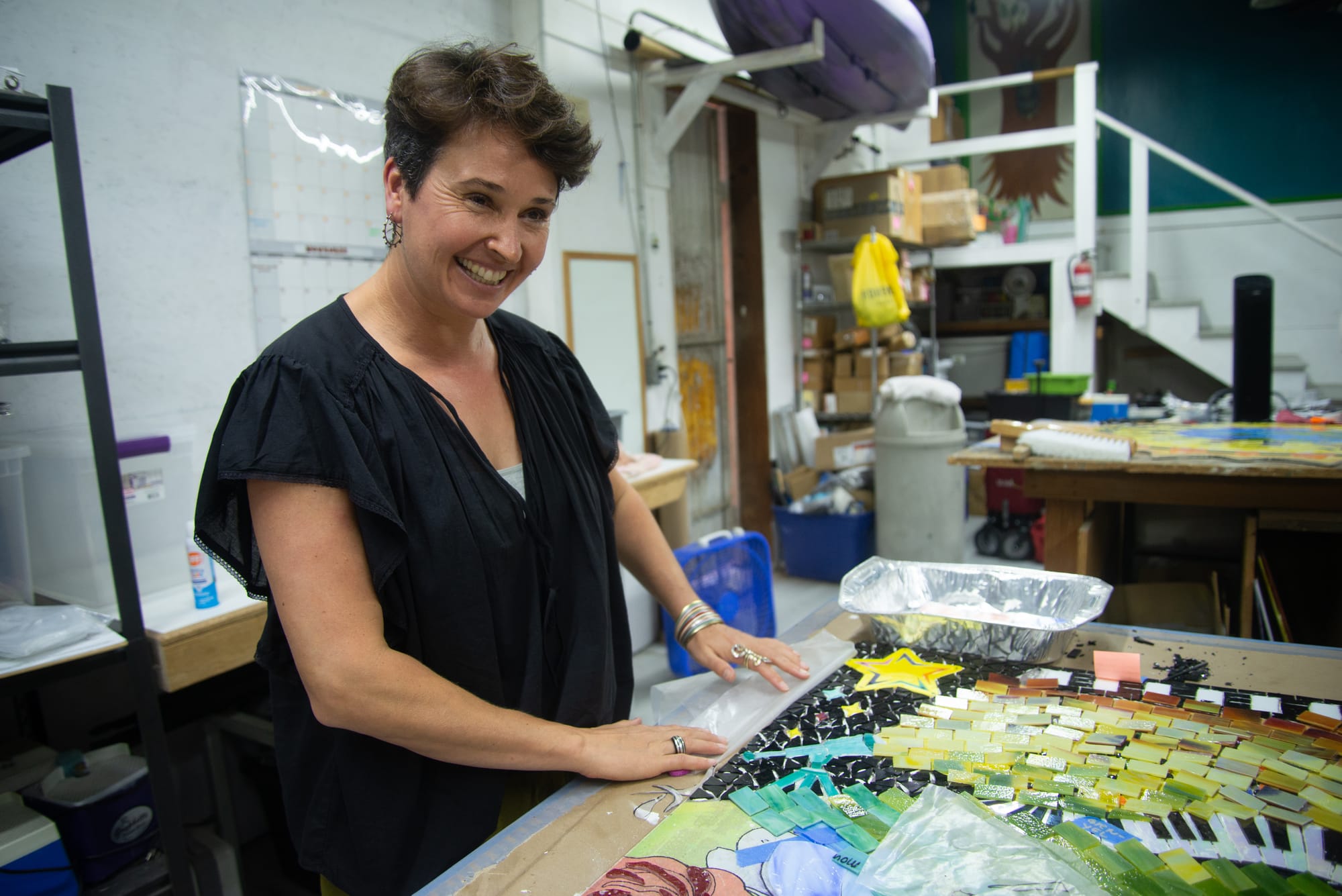 A woman with short brown hair and a black shirt stands next to a table with pieces of tile laid out as part of a mosaic
