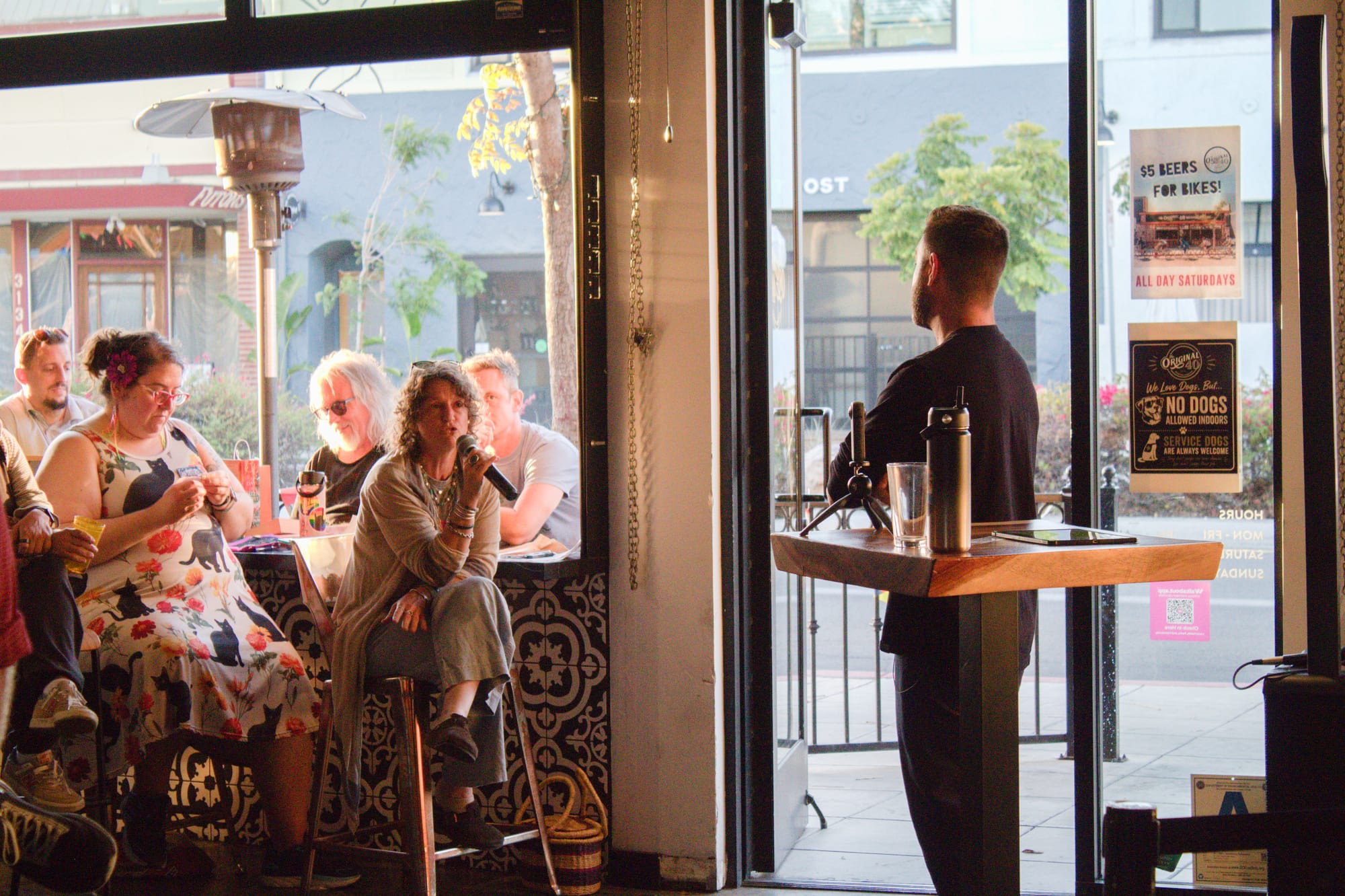 In a bar, a woman sits on a stool while talking into a mic toward a man standing and facing her.